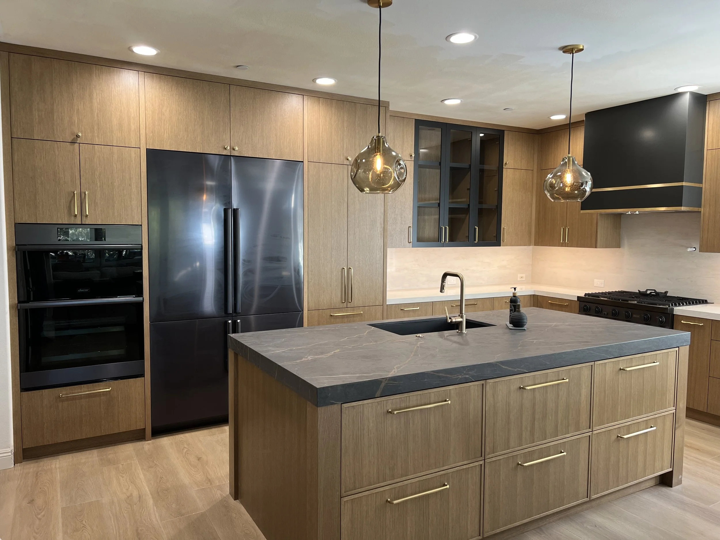 Luxury kitchen design with rift white oak cabinets, dacor graphite appliances, dekton island counter, custom designed hood, statement satin gold cabinet hardware, black upper cabinet with glass doors, led under counter lights 