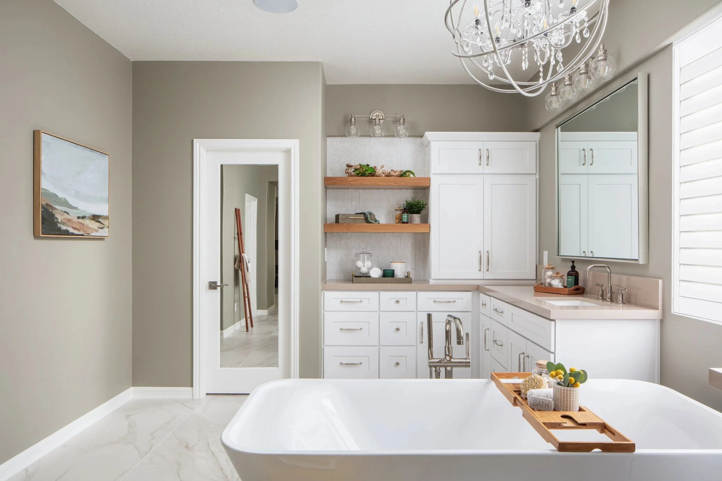 Spa like bathroom remodel with a white bathtub, white cabinetry, and a large mirror. The decor includes wooden accents, a chandelier, and a small tray with bath accessories and plants.