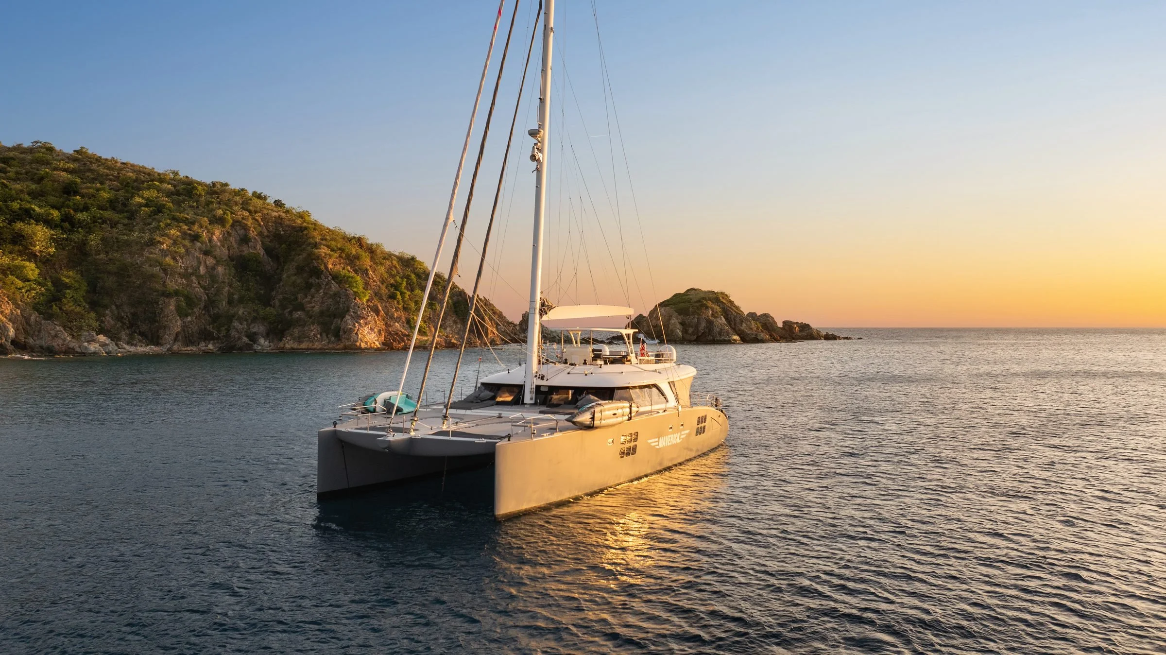 MAVERICK at anchor in late golden light beside a rocky green headland, BVI.