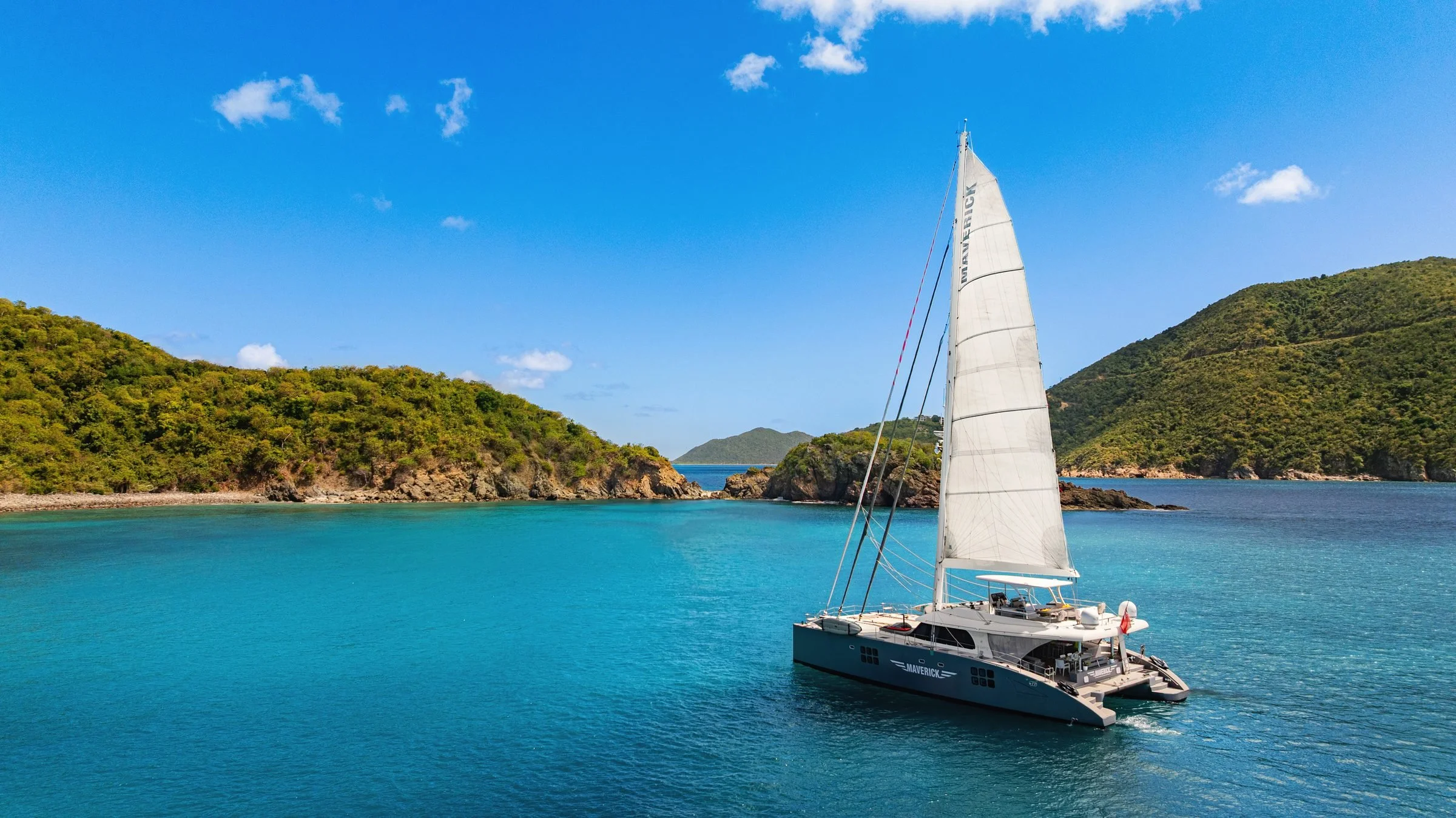 MAVERICK under her mainsail in turquoise water between rocky green headlands, BVI.