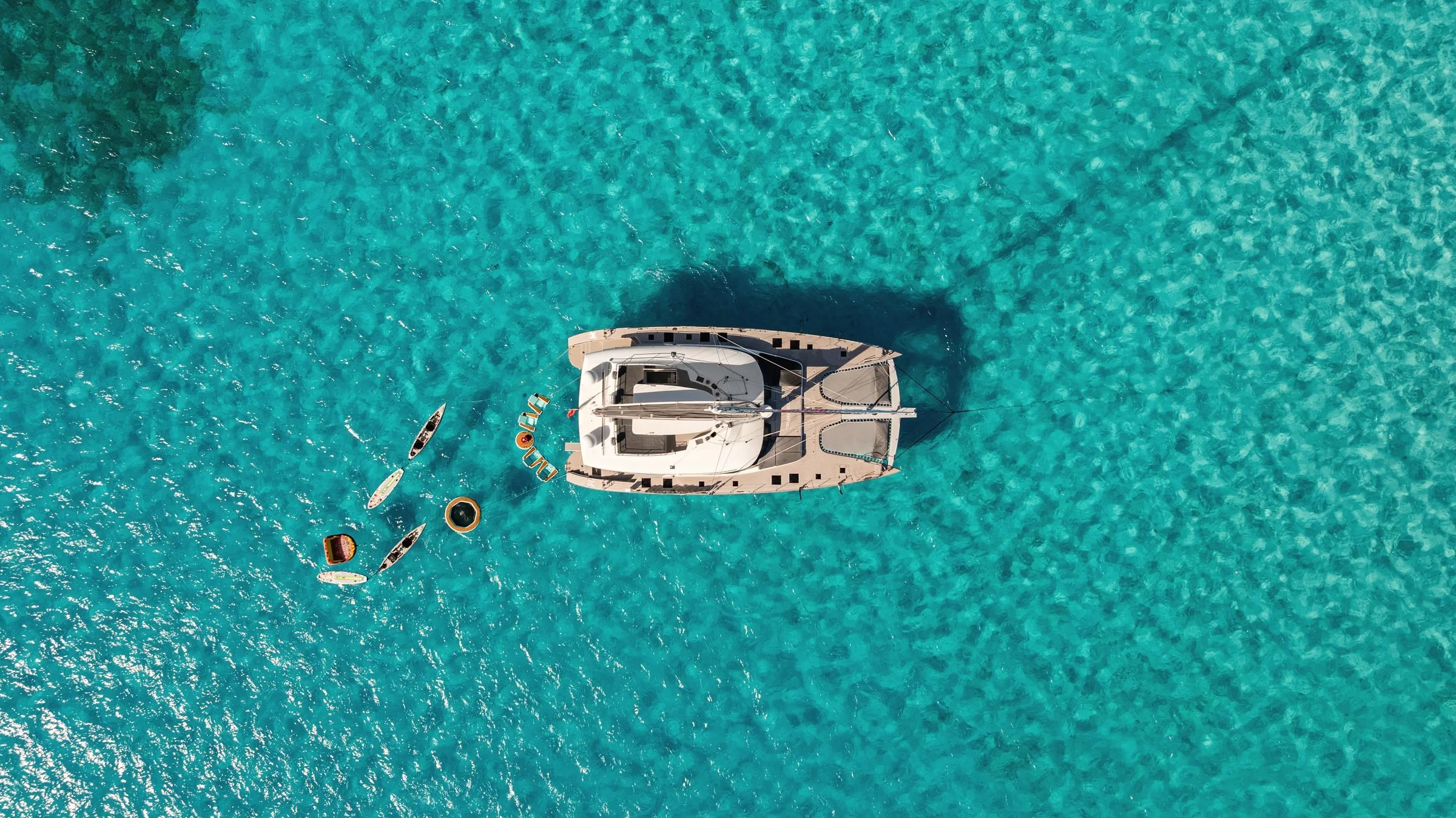MAVERICK at anchor seen from directly above, with kayaks, paddleboards and floats behind her in turquoise water, BVI.