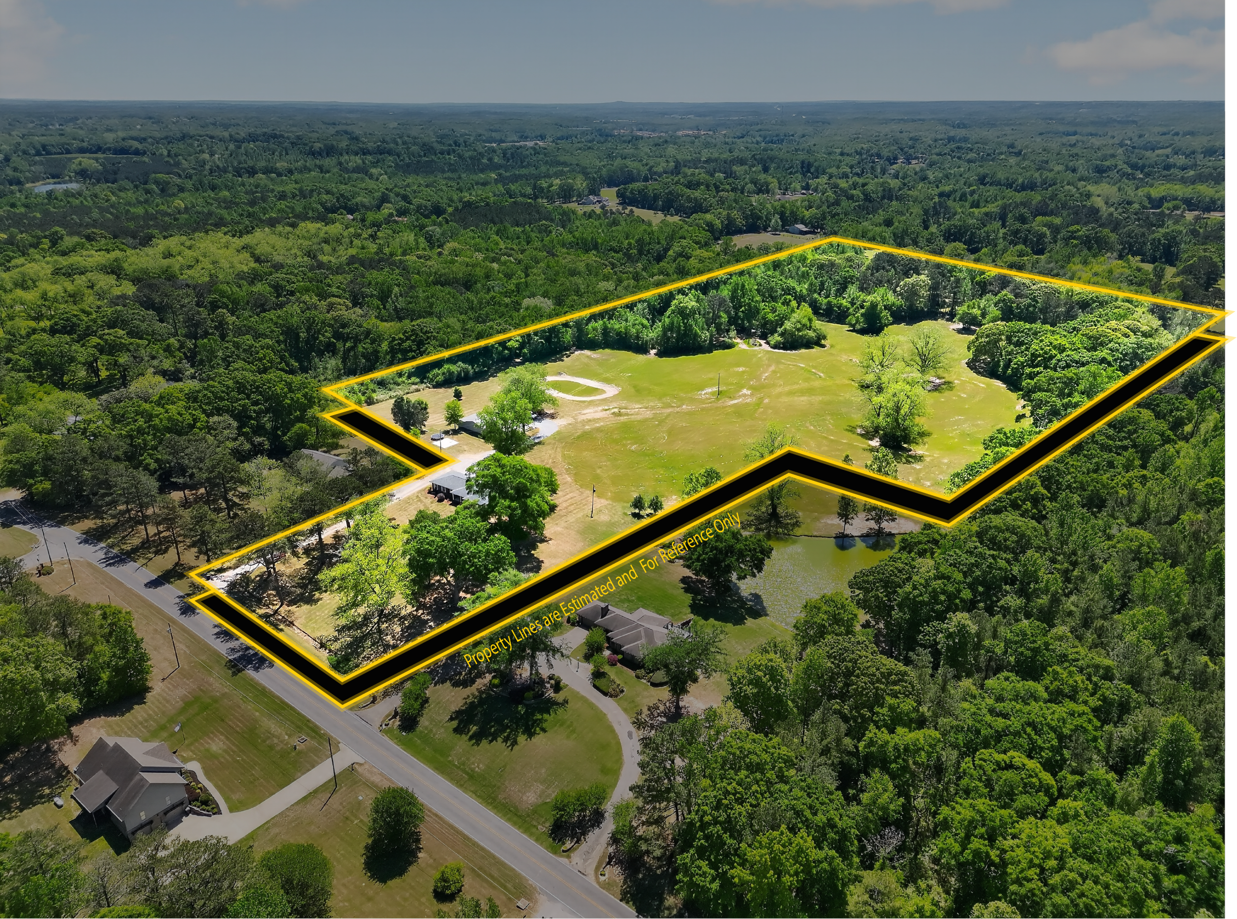 Aerial view of a large property outlined with yellow property lines in a lush green area with trees and open grassy spaces. Houses and a road are visible in the foreground.