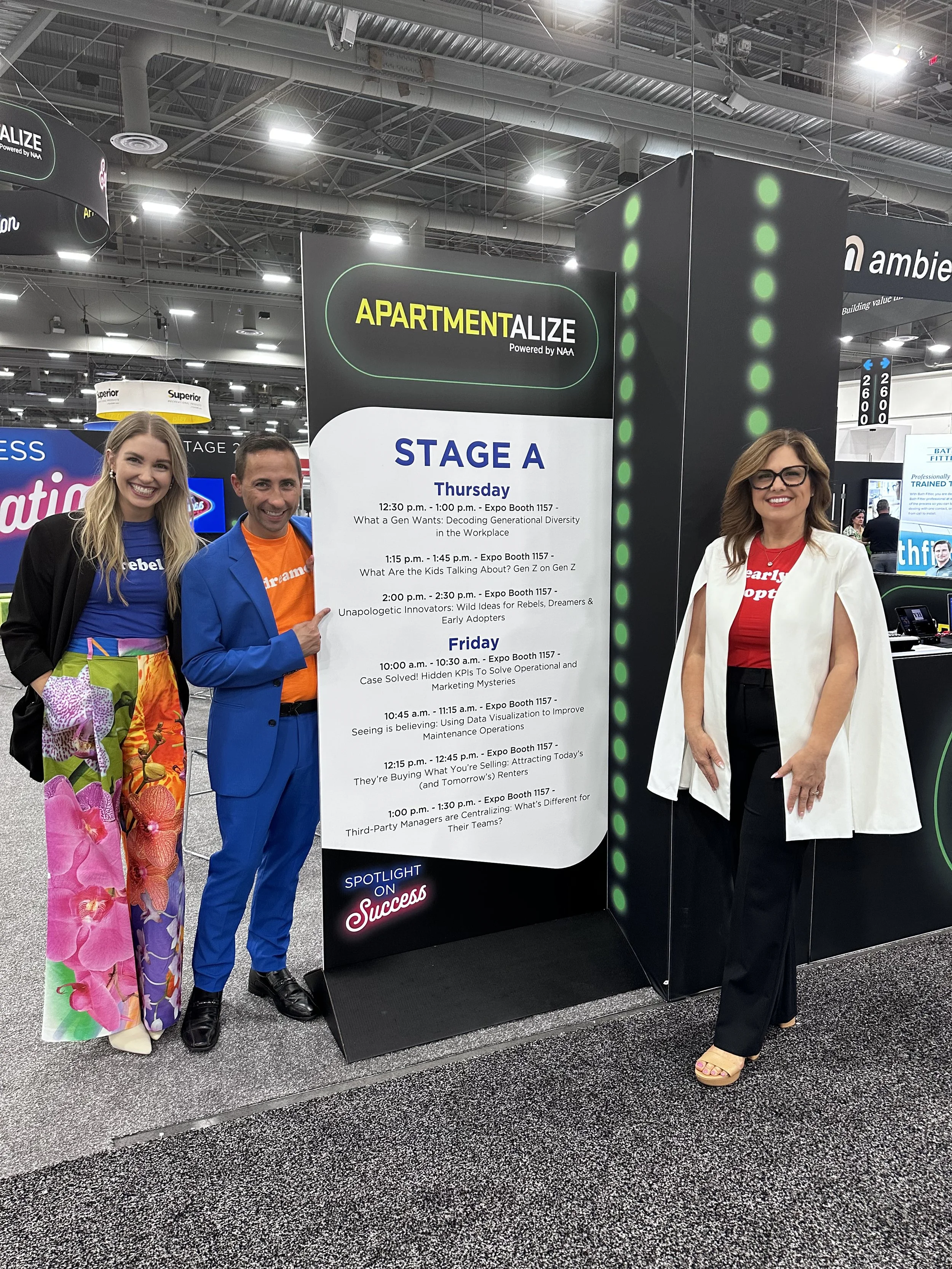 Three presenters standing next to a schedule board at a conference, with stage information for 'Apartmentalize' on Thursday and Friday.