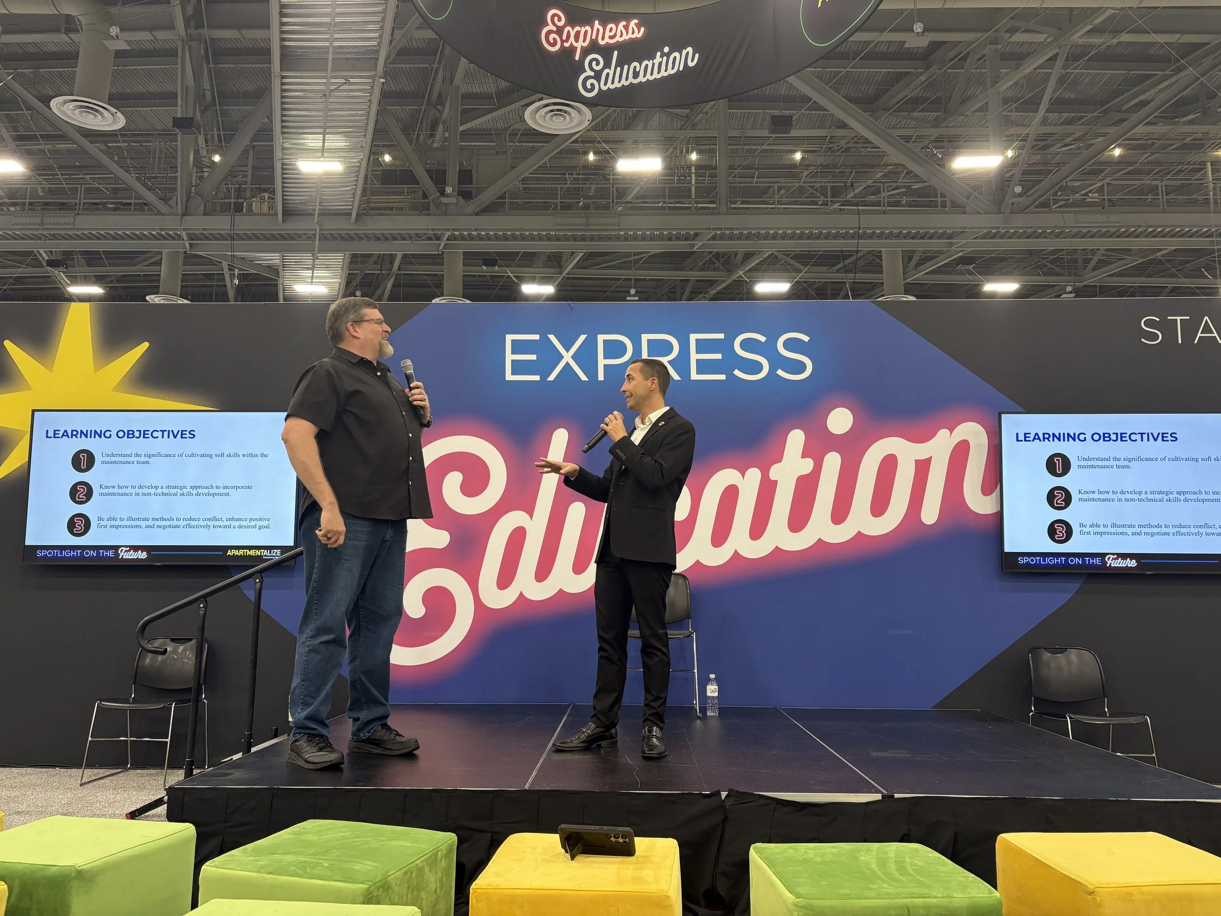 Two men are standing on a stage engaged in conversation at a conference. One is holding a microphone, and the other is gesturing with his hand. There are two screens on either side of the stage displaying learning objectives. The background features