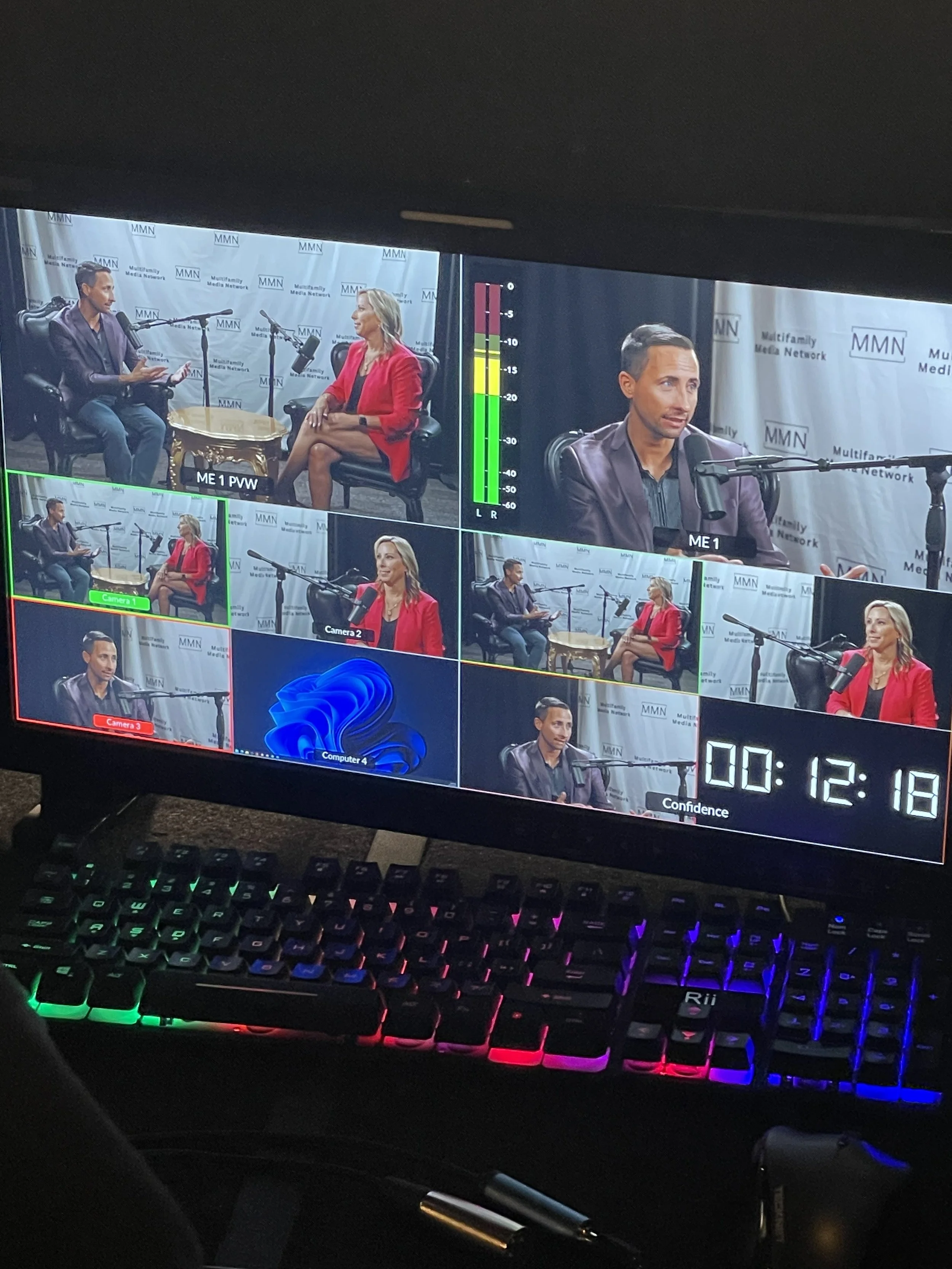 A multi-camera view of a recorded interview in a radio studio featuring two people, a man and a woman, sitting in front of microphones. The background shows a banner with 'Multifamily Media Network' and 'MMN'. The monitor displays various camera angl