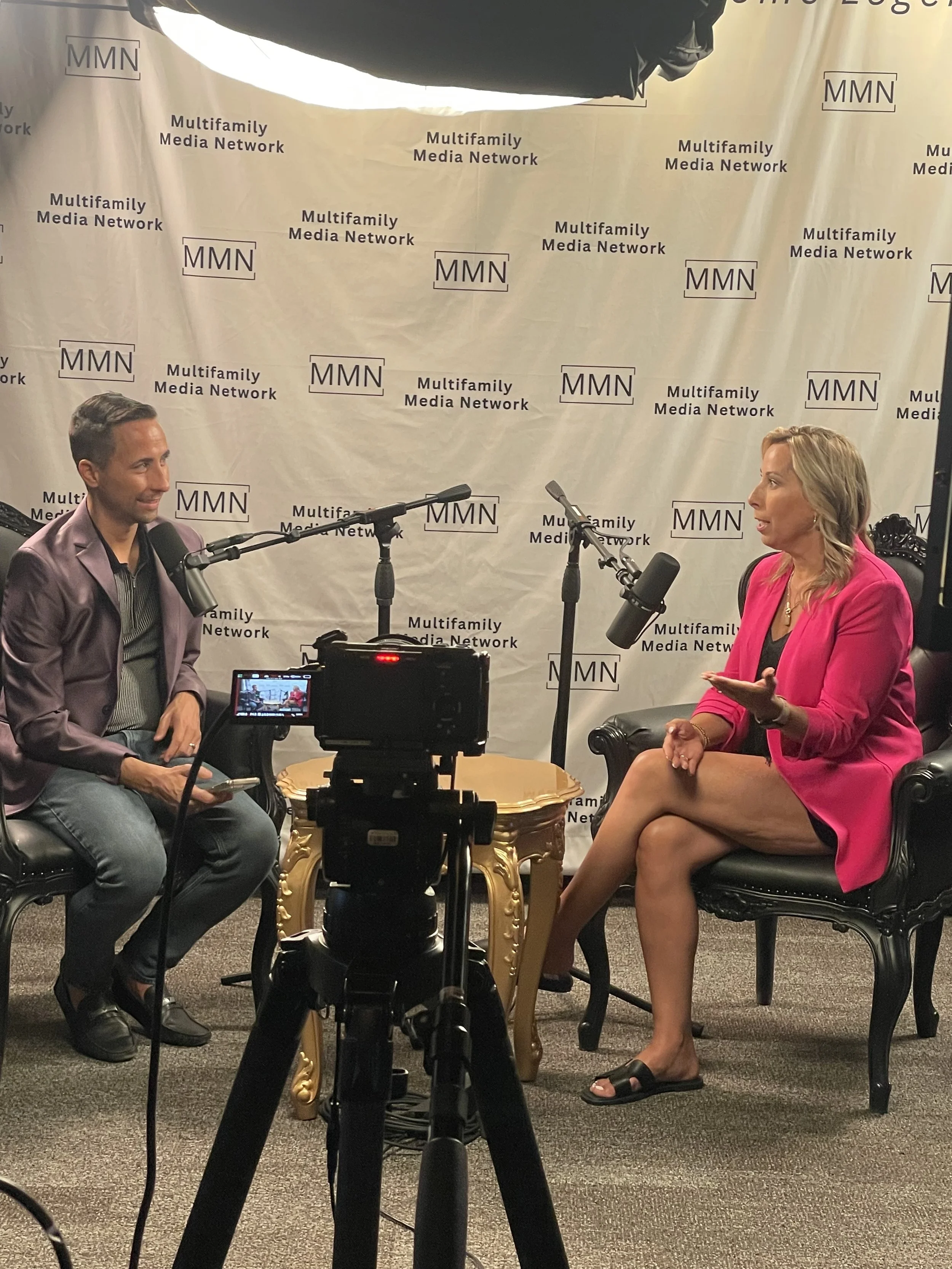 A woman and a man are sitting in black chairs on a set during a recorded interview. The woman is wearing a pink blazer, black dress, and sandals, and the man is in a purple blazer and gray pants. They are speaking into microphones, with a camera cent