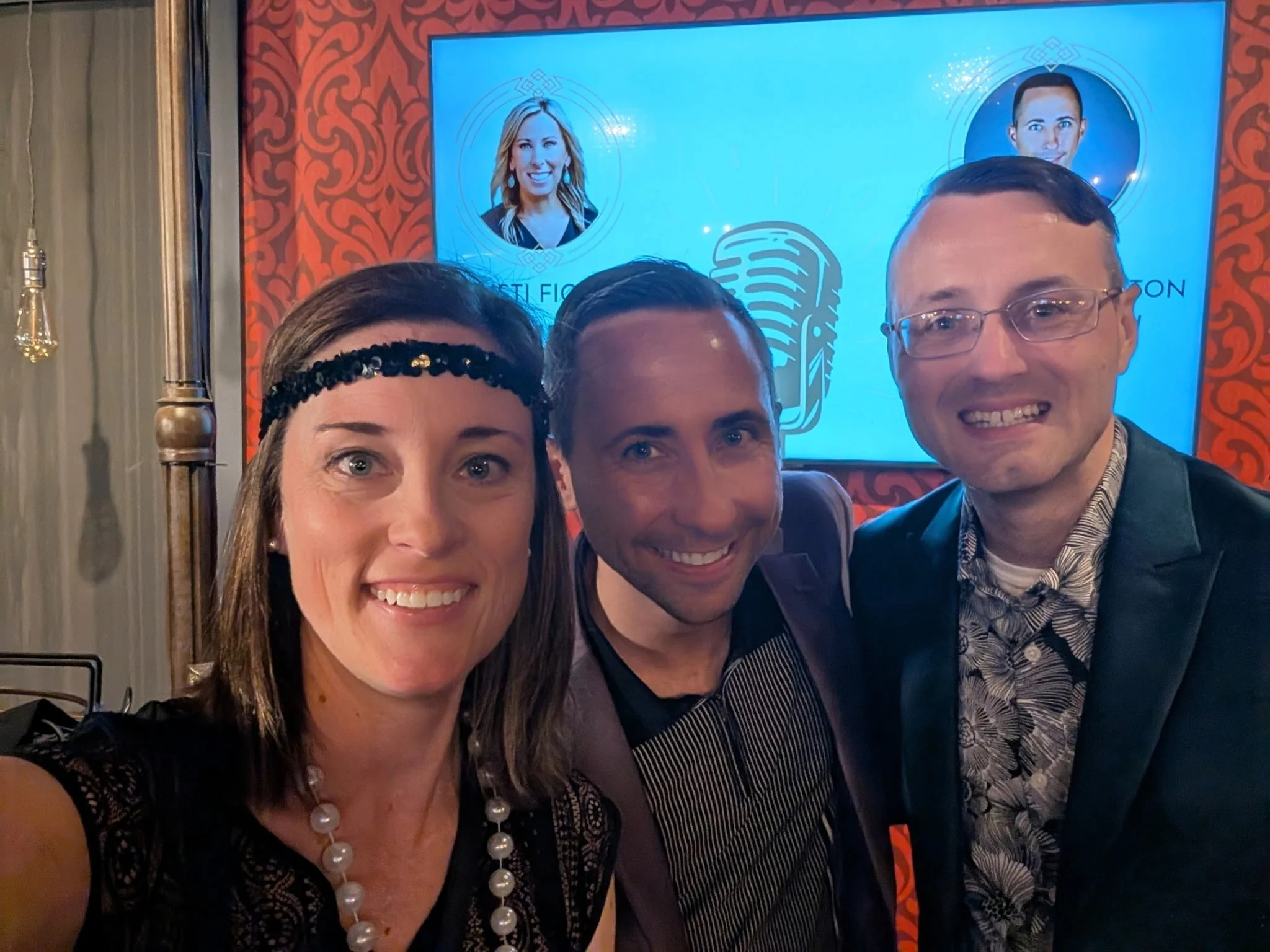 Three people taking a selfie in front of a blue screen with photos of a woman and a man, and a microphone graphic, at a formal event.