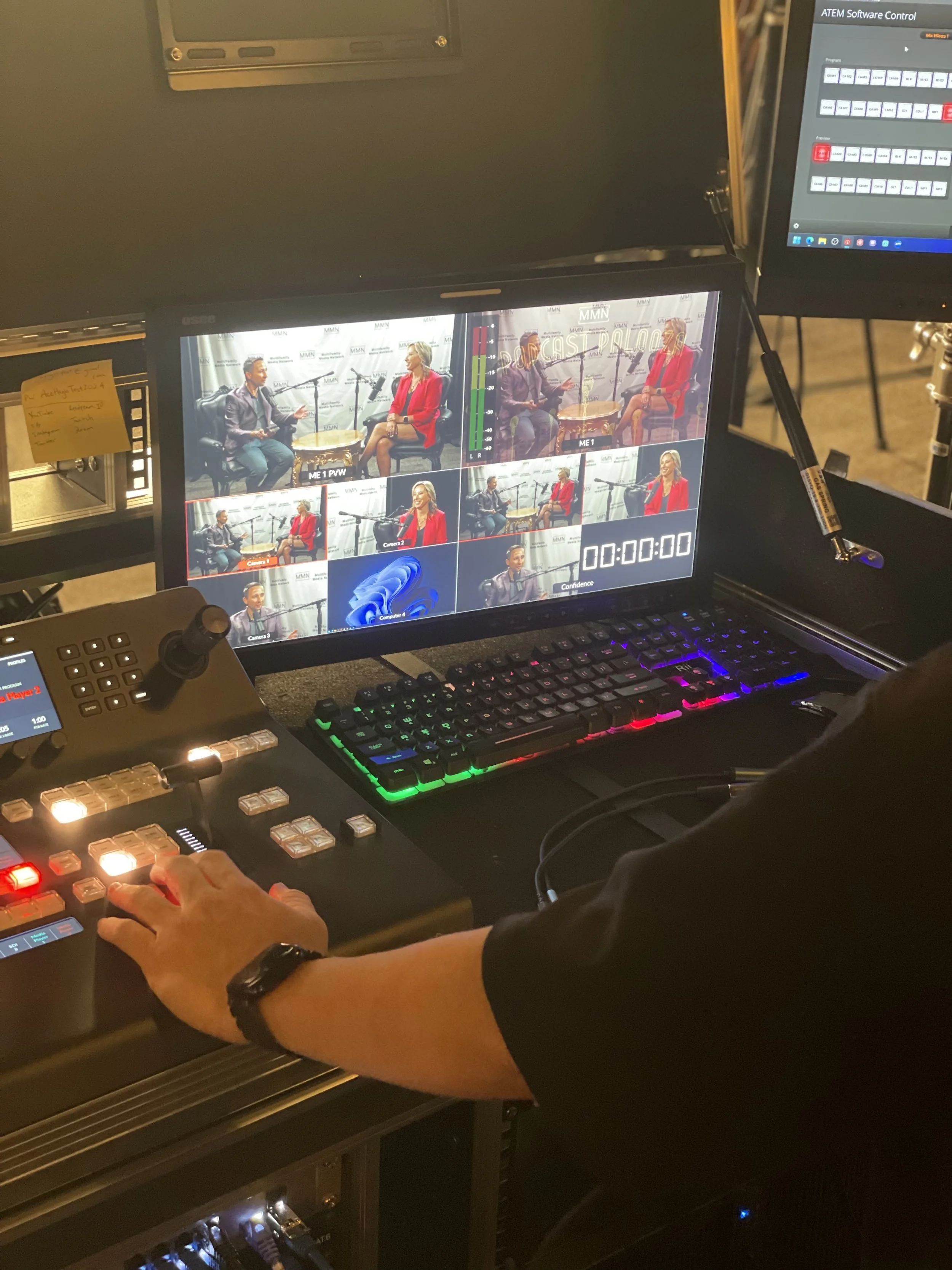 Video control room with a monitor showing nine small frames of an interview with two women, a person operating a soundboard, a keyboard with multicolored backlighting, and another monitor displaying software control.