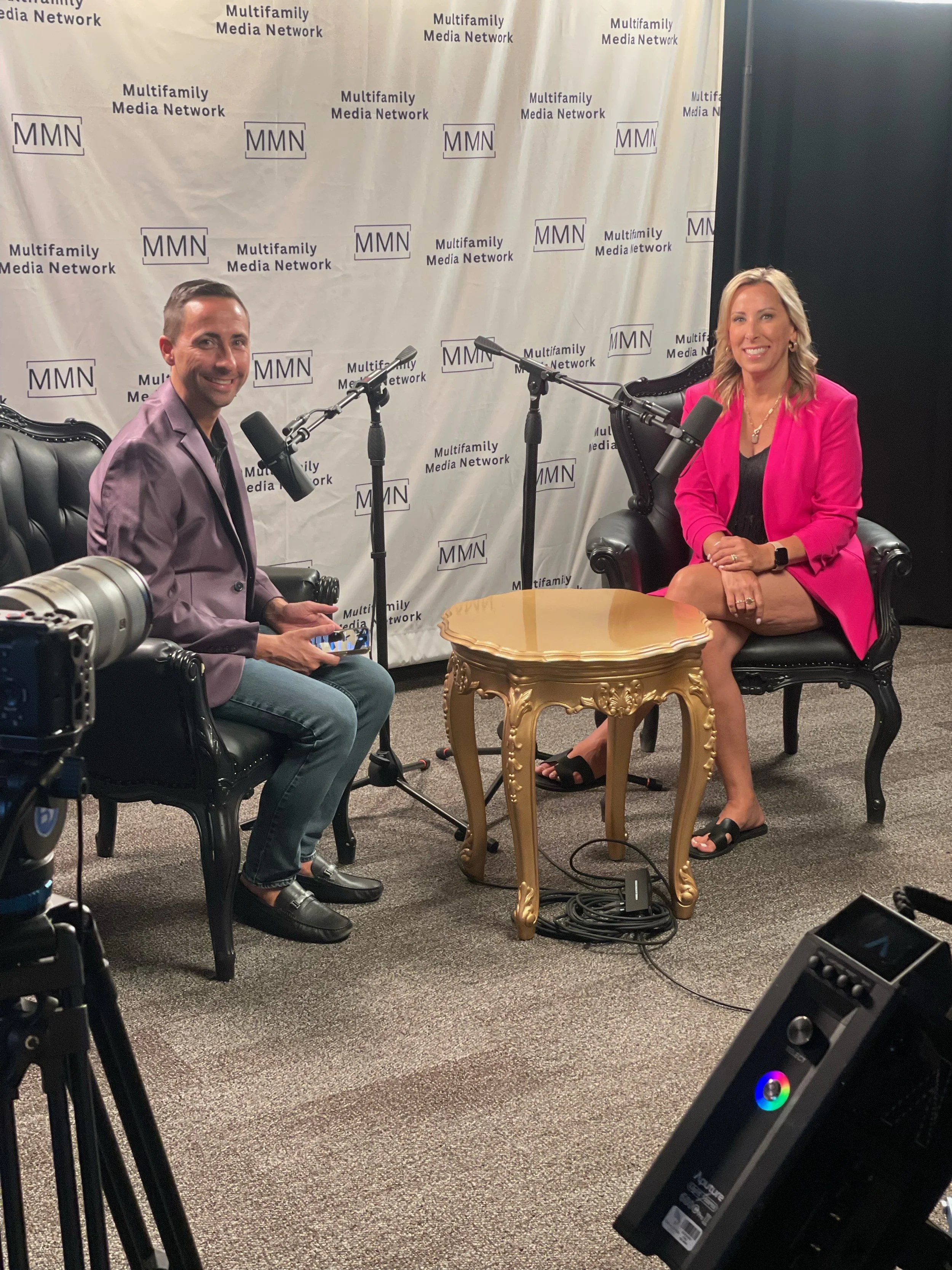 Two people sitting in black ornate chairs in front of a backdrop with multiple "Multifamily Media Network" logos, microphones positioned between them, and a gold-colored small table in the middle. The woman on the right is wearing a bright pink blaze