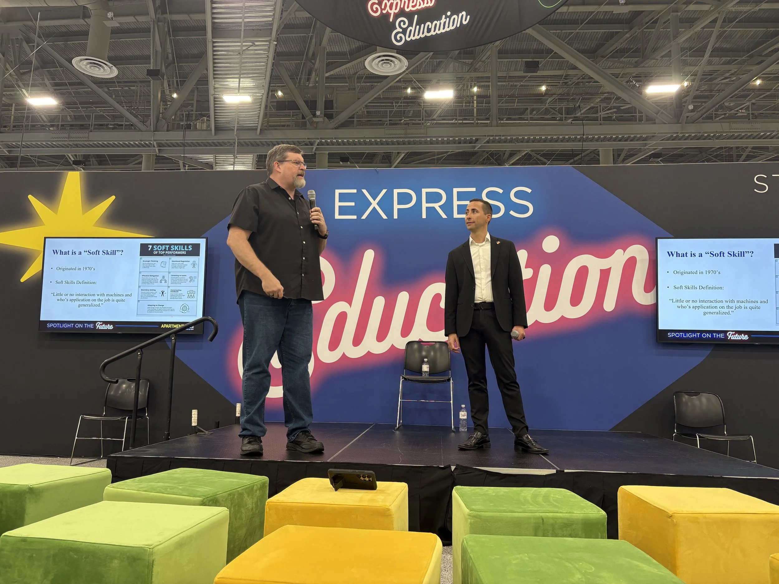 Two men are standing on a stage with a large sign behind them that reads 'Express Education'. One man is speaking into a microphone and the other is looking at him. There are two screens on either side of the stage displaying slides, and colorful sea