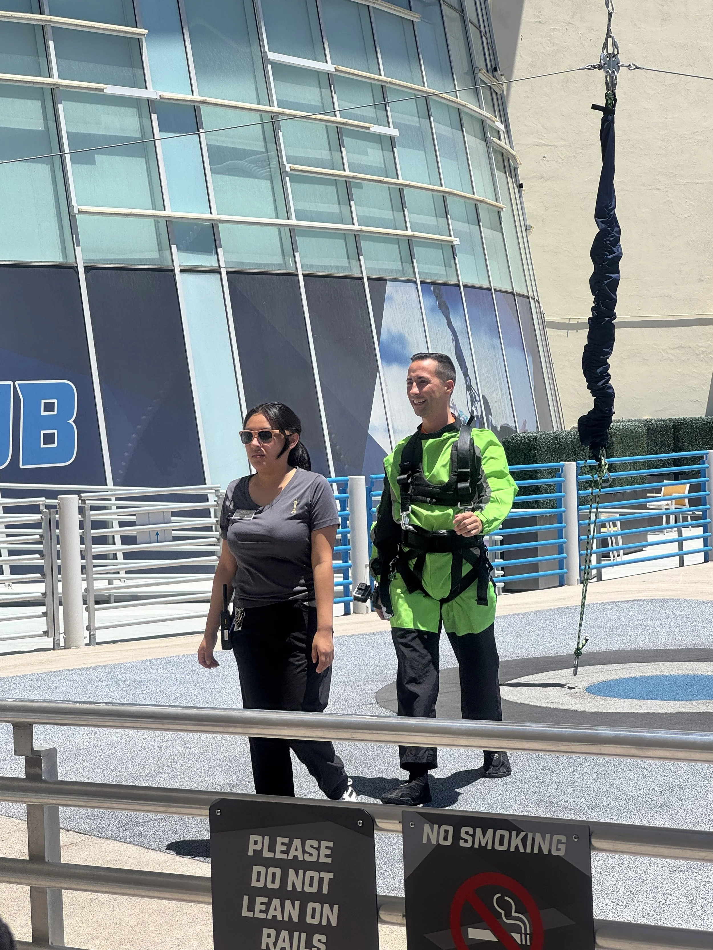 Two people, a woman and a man, standing outside near a sign that says, "Please do not lean on rails" and "No smoking." The man is wearing a bright green and black suit with harnesses, and the woman is wearing a gray t-shirt, sunglasses, and dark pant