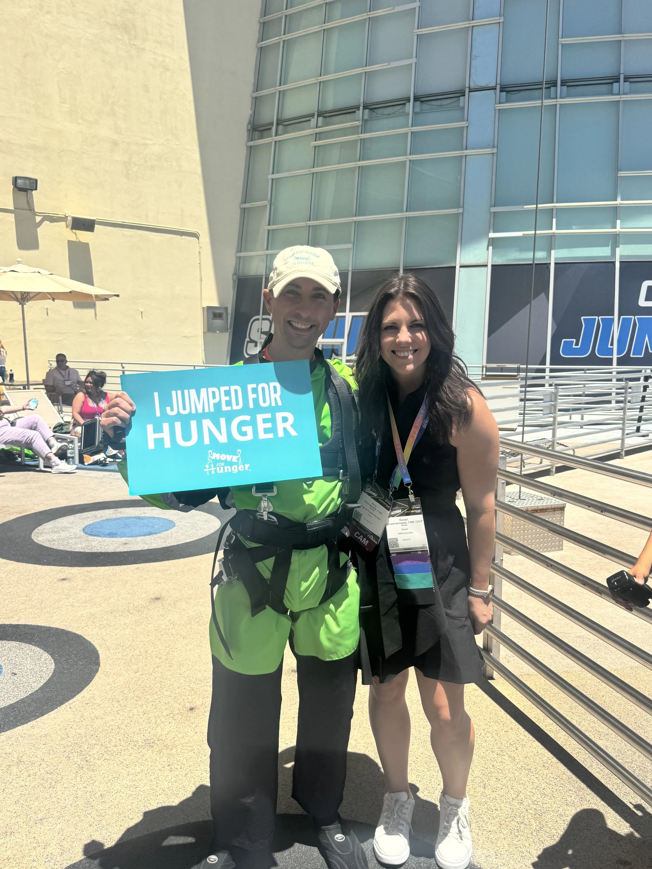 Two women standing outside at a crowd event, one holding a sign that says "I JUMPED FOR HUNGER." The woman on the left is wearing a bright green jacket, black pants, and a white cap, while the woman on the right is dressed in a black dress with white