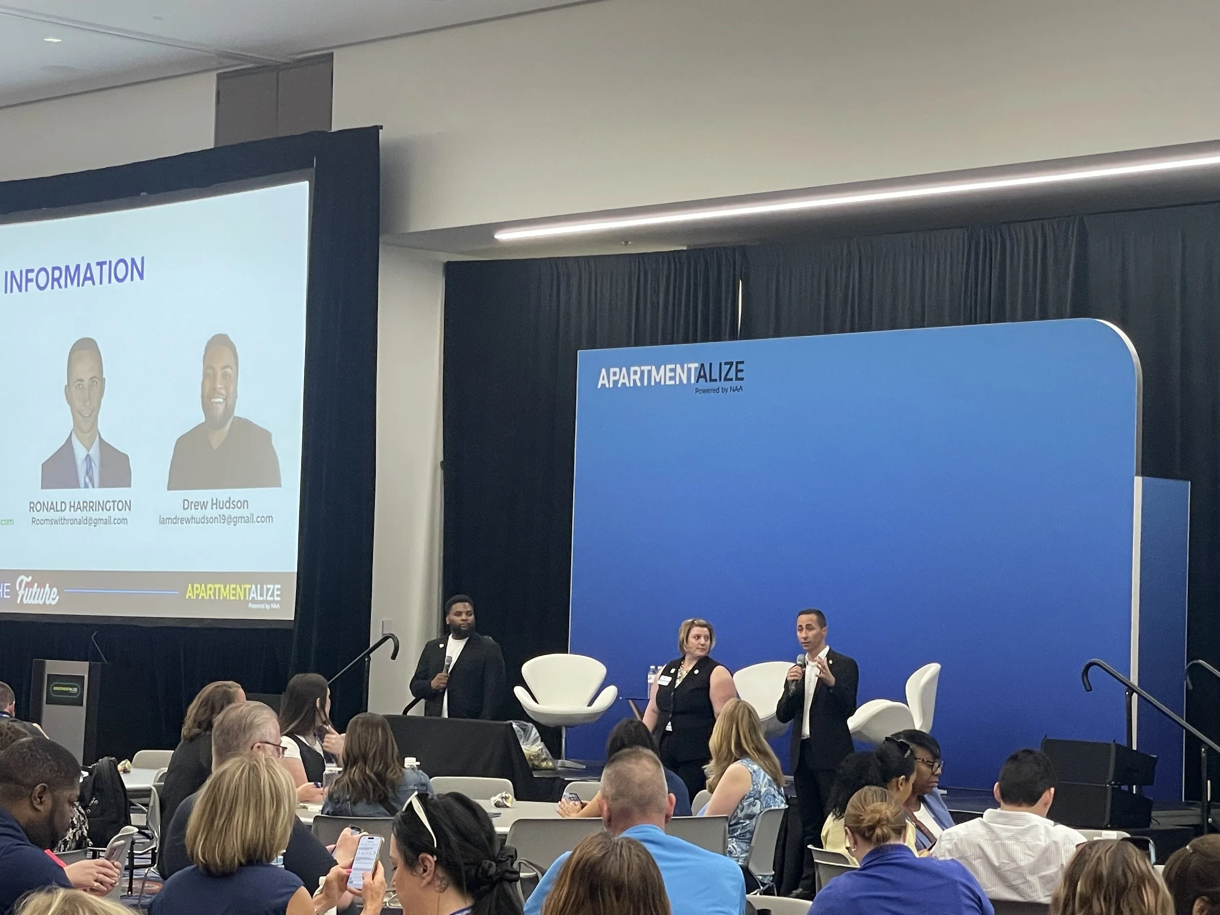 A conference or panel discussion with two women and one man on stage, with a large audience watching, and a presentation screen displaying two speakers' photos and info.