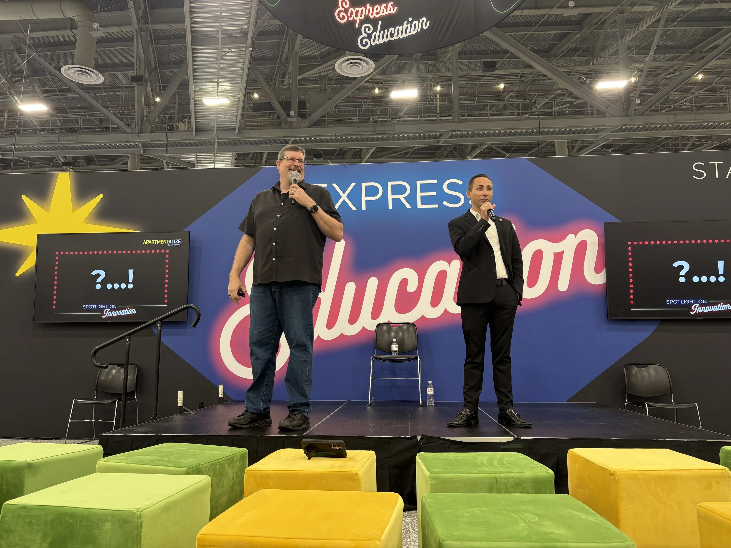 Two men standing on a stage with a large sign that reads 'EXPRESS' and a backdrop with 'Celebration' in stylized text. One man is wearing a black short-sleeve shirt and jeans, holding a microphone, while the other is in a black suit and tie, also hol