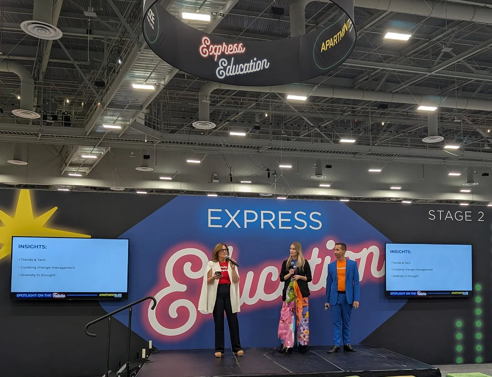 Three people on stage at a conference, standing in front of a backdrop that reads 'EXPRESS Education' with a yellow star graphic. Two screens on either side display insights on trends, tech, change management, and diversity in thought. The woman on t