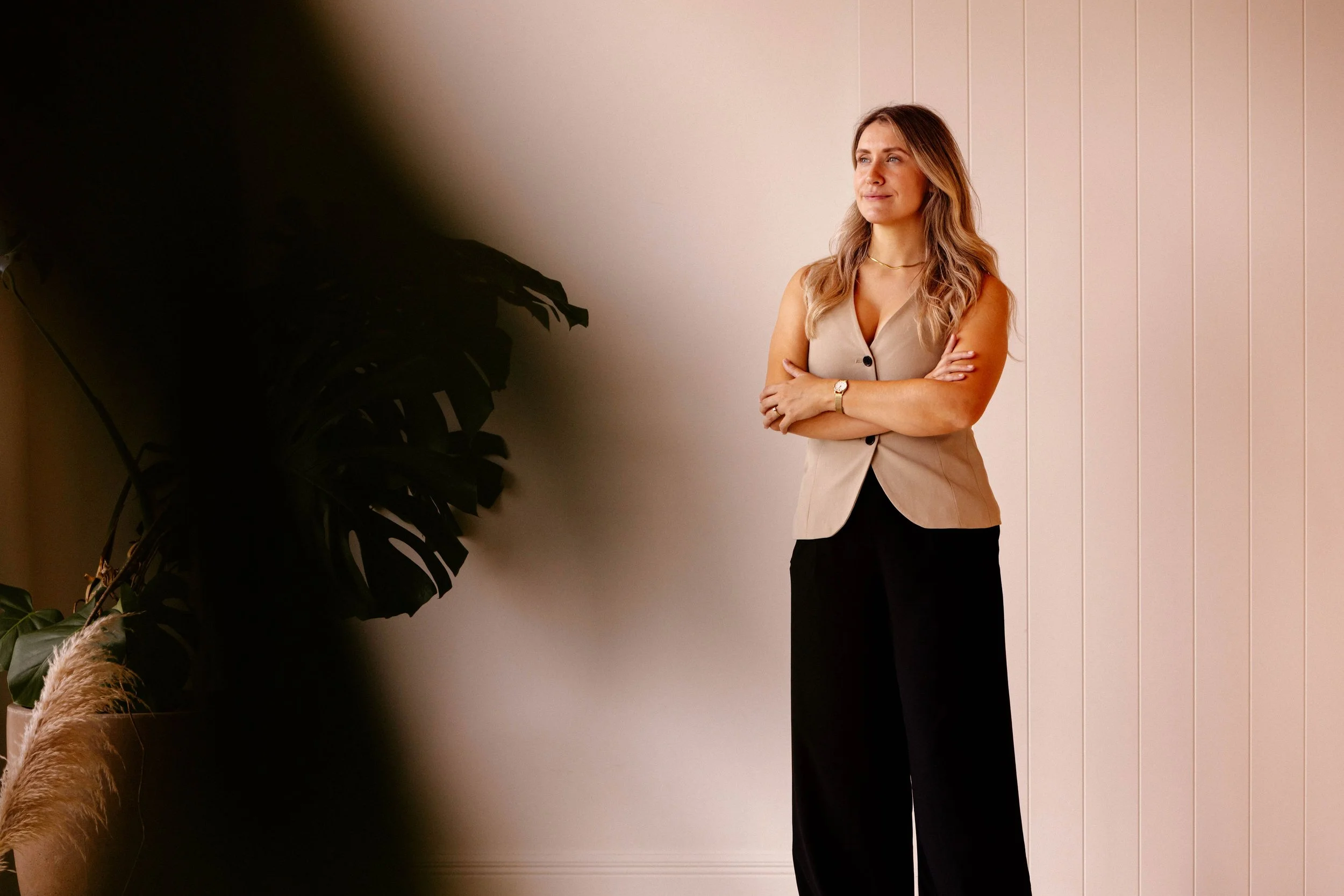 Woman in a beige vest and black pants standing with arms crossed, looking to the side, near a large plant indoors.