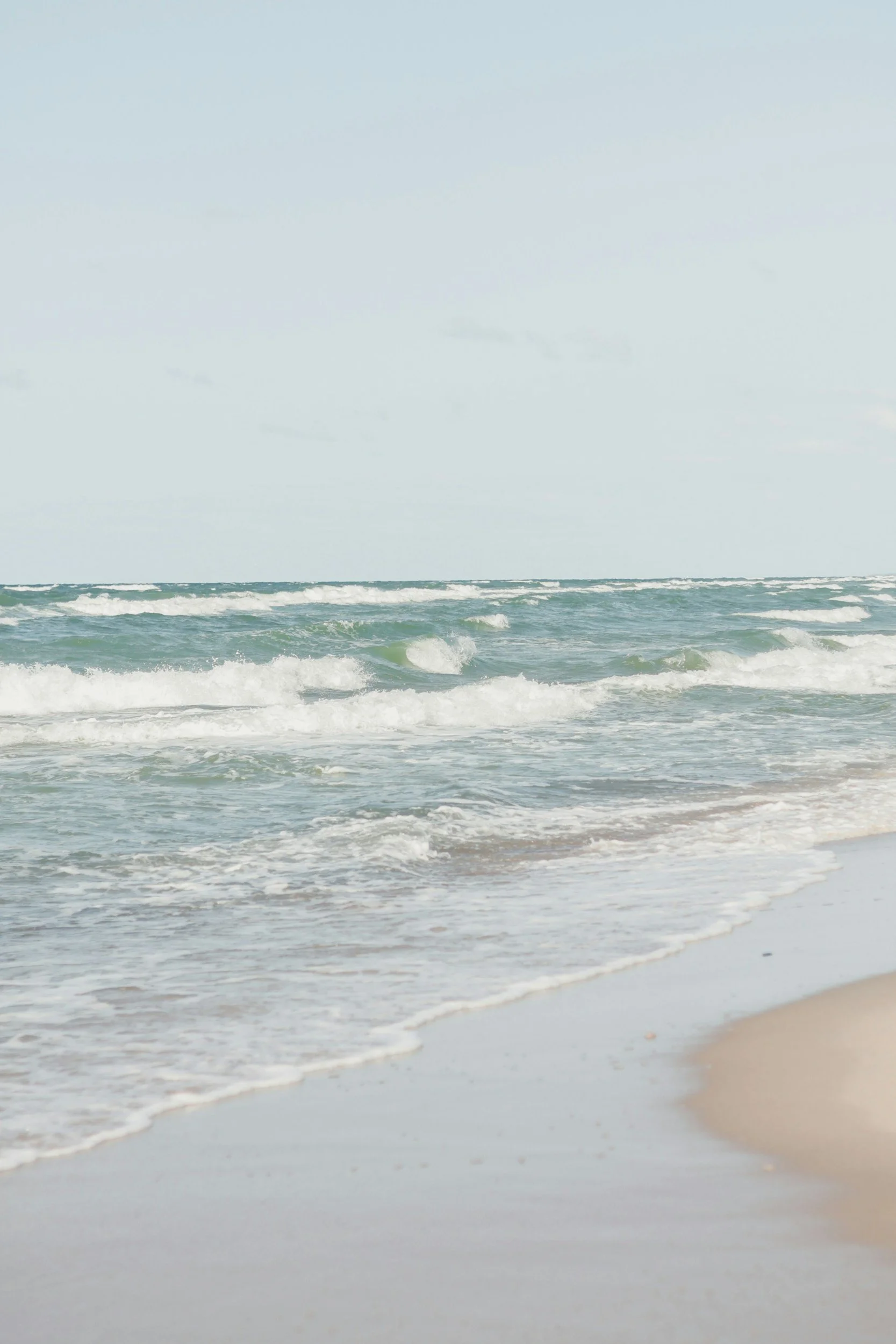 A view of the ocean with gentle waves approaching the sandy shore, under a slightly cloudy sky.