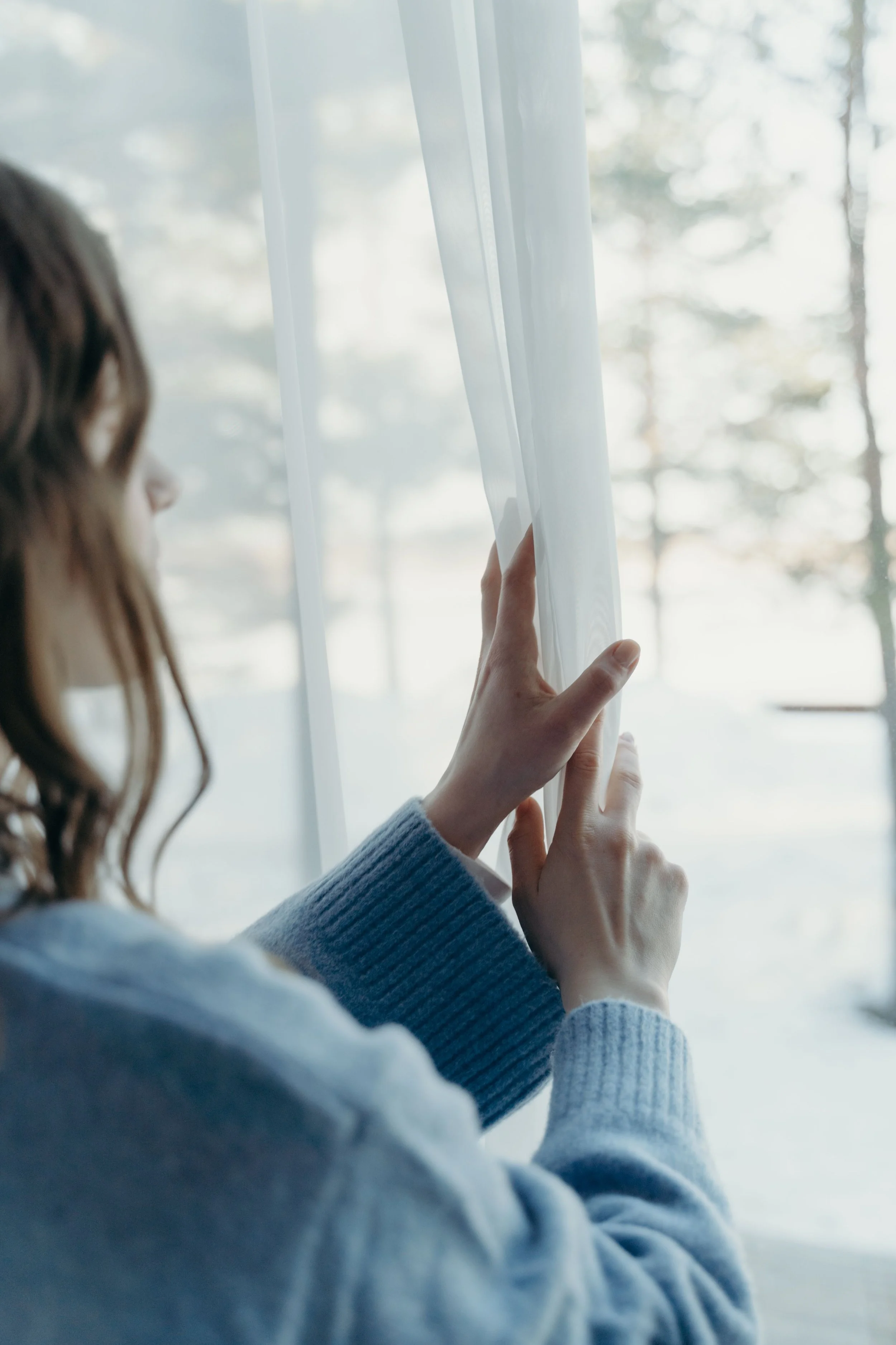 A woman with wavy hair, viewed from the side, is touching a sheer white curtain near a window. Behind her, outdoors, there are blurred trees and natural light coming in.