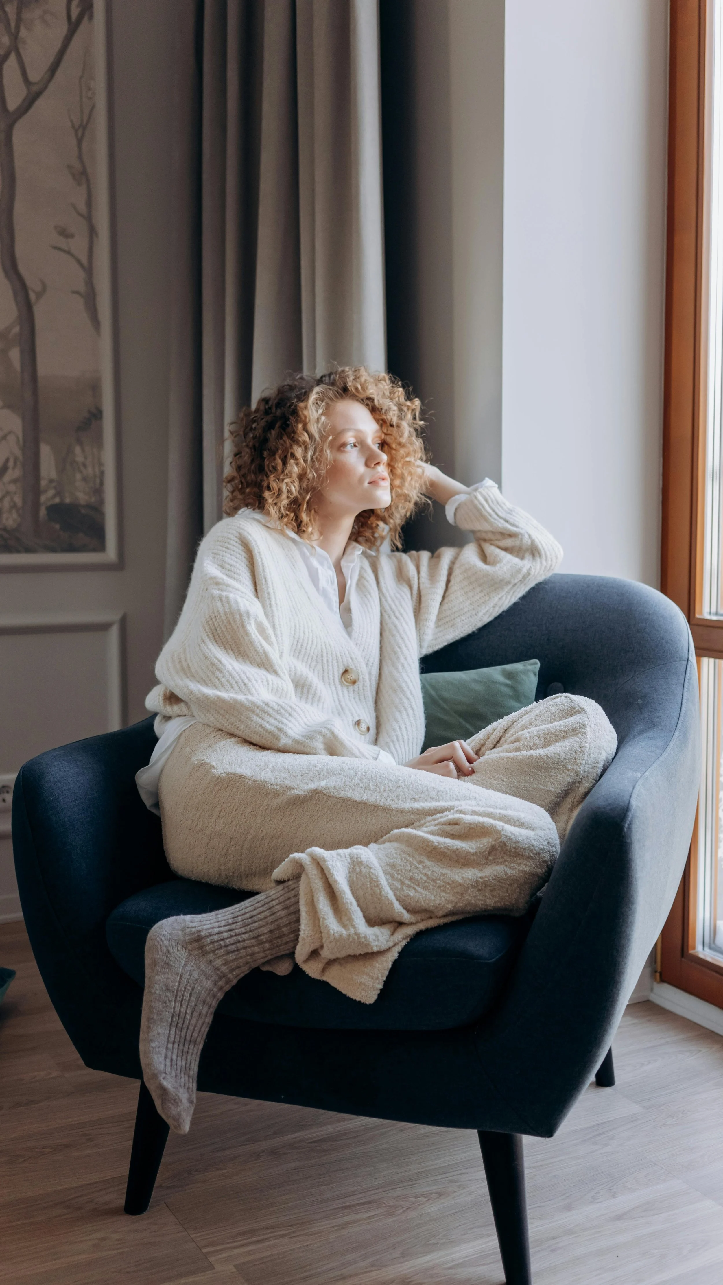 A young woman with curly red hair sitting on a dark blue armchair by a window, looking outside thoughtfully. She is dressed in cozy, cream-colored loungewear.