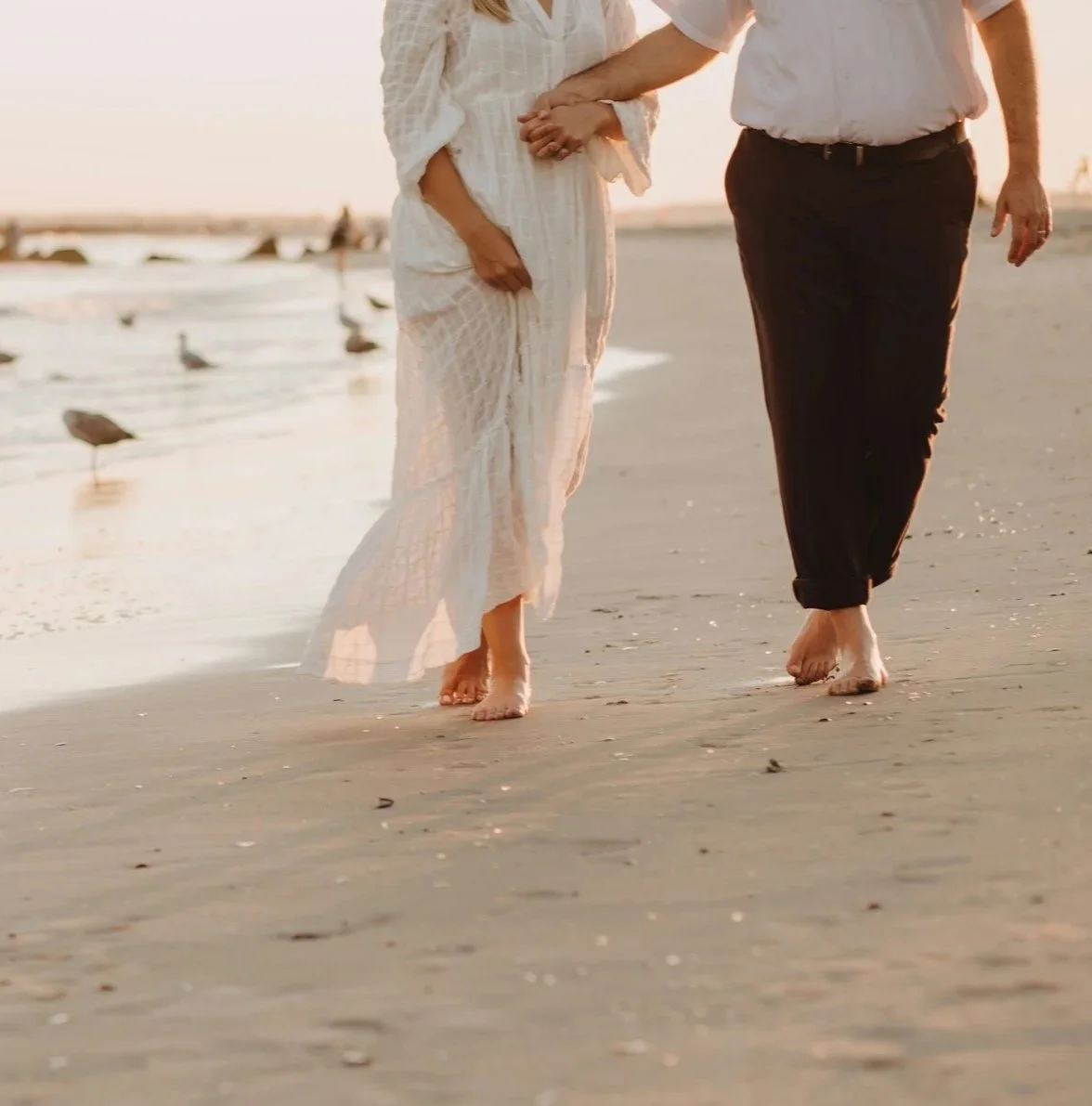 A couple holding hands and walking barefoot on the beach during sunset.