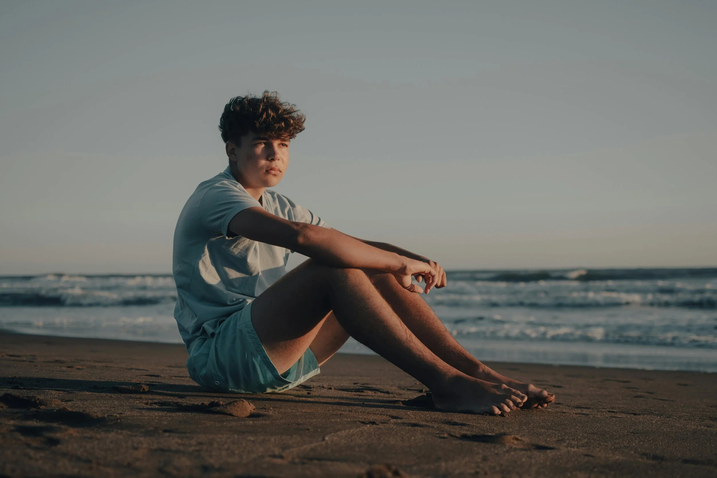 A young man with curly hair sitting on the beach, looking thoughtfully at the ocean during sunset.
