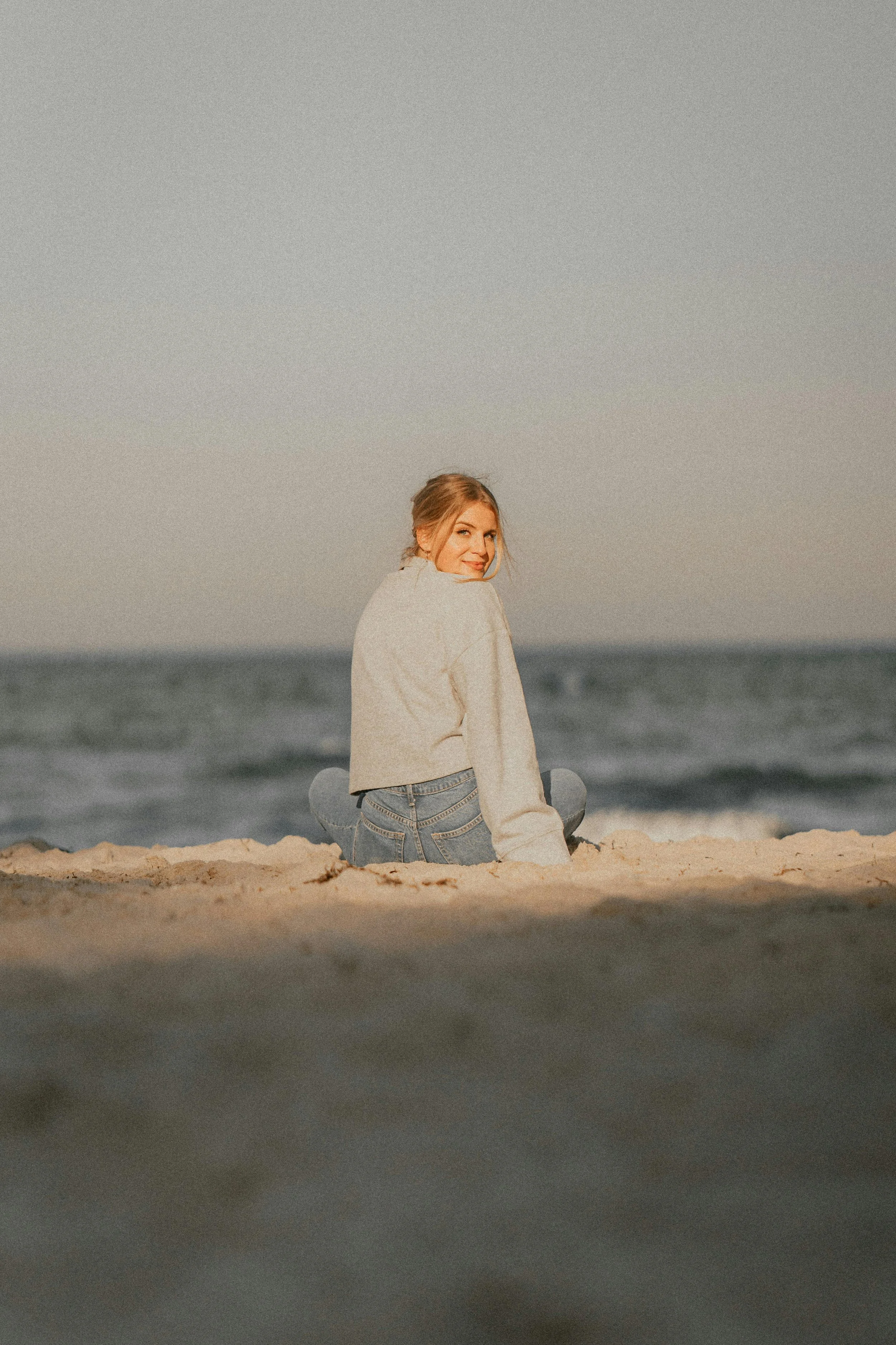 A young woman with blonde hair sitting on the beach with waves in the background, looking over her shoulder and smiling.
