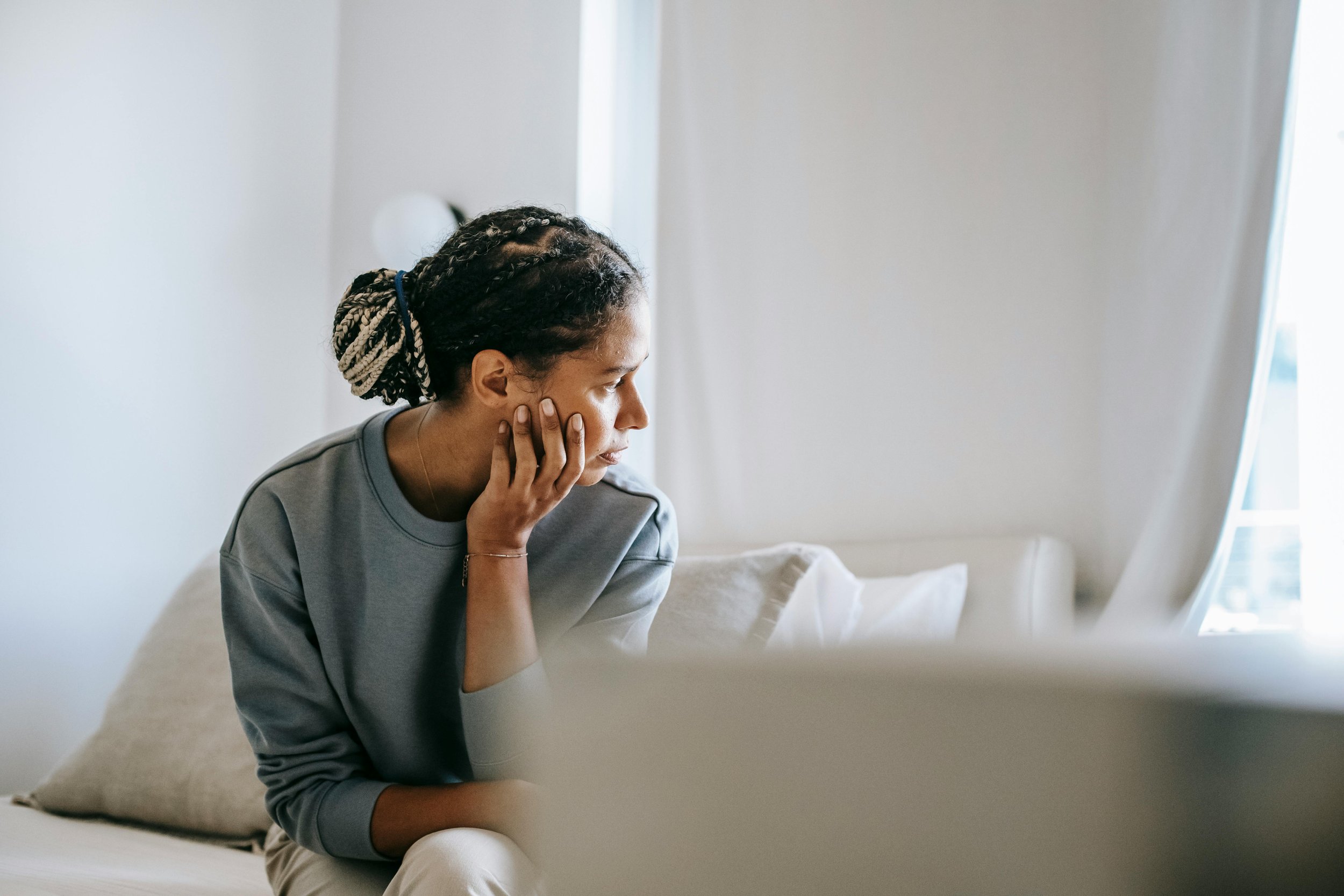 A woman with braided hair sitting on a bed, resting her head on her hand, looking at a mirror in a bright room.
