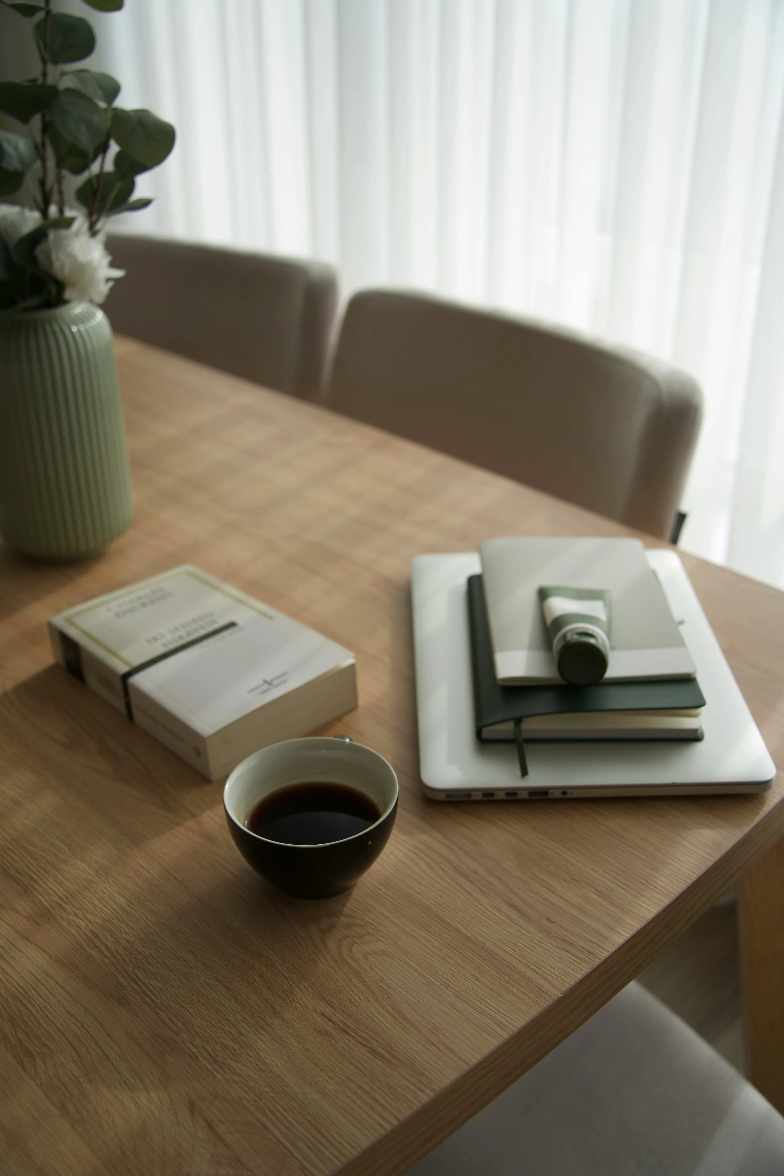 A wooden table with a black coffee cup, a white closed box, a stack of books with a gray pen and a white tablet on top, a light green vase with white flowers and green leaves, and a white curtain in the background.