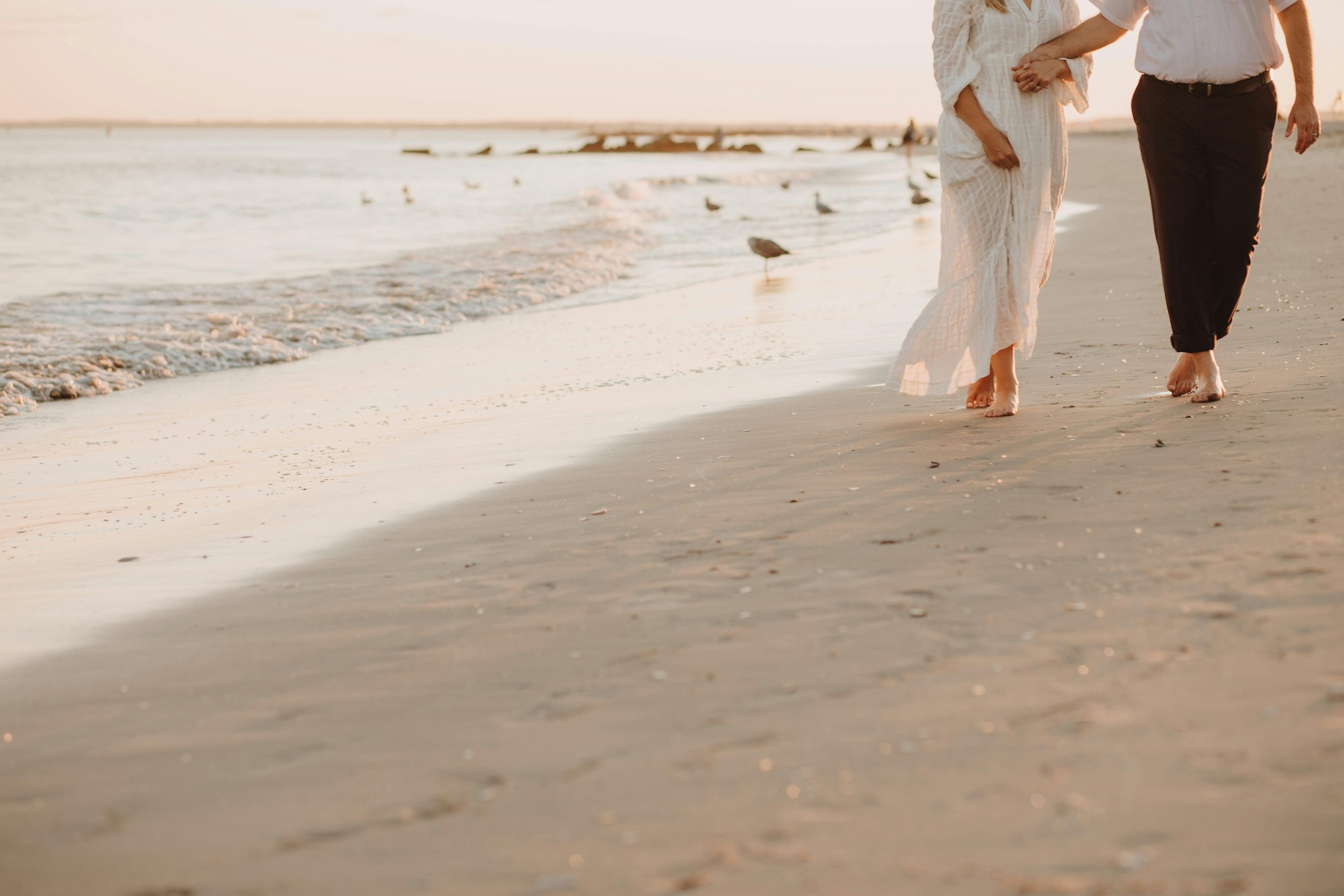 People walking barefoot along the shoreline at sunset with seagulls nearby.