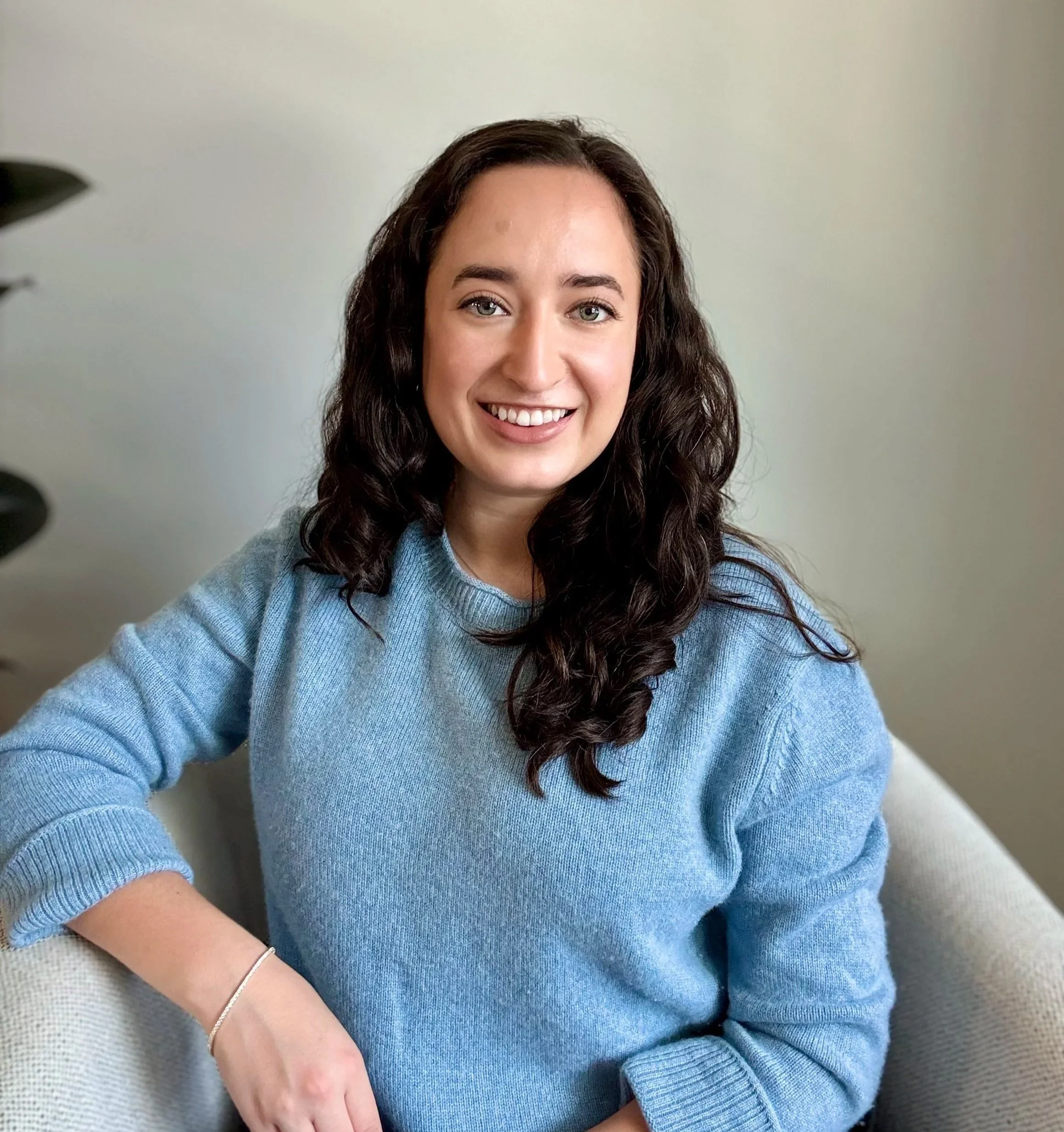 A woman with dark curly hair smiling, wearing a light blue sweater, sitting on a beige sofa in a room with a light-colored wall and a plant partially visible on the left.