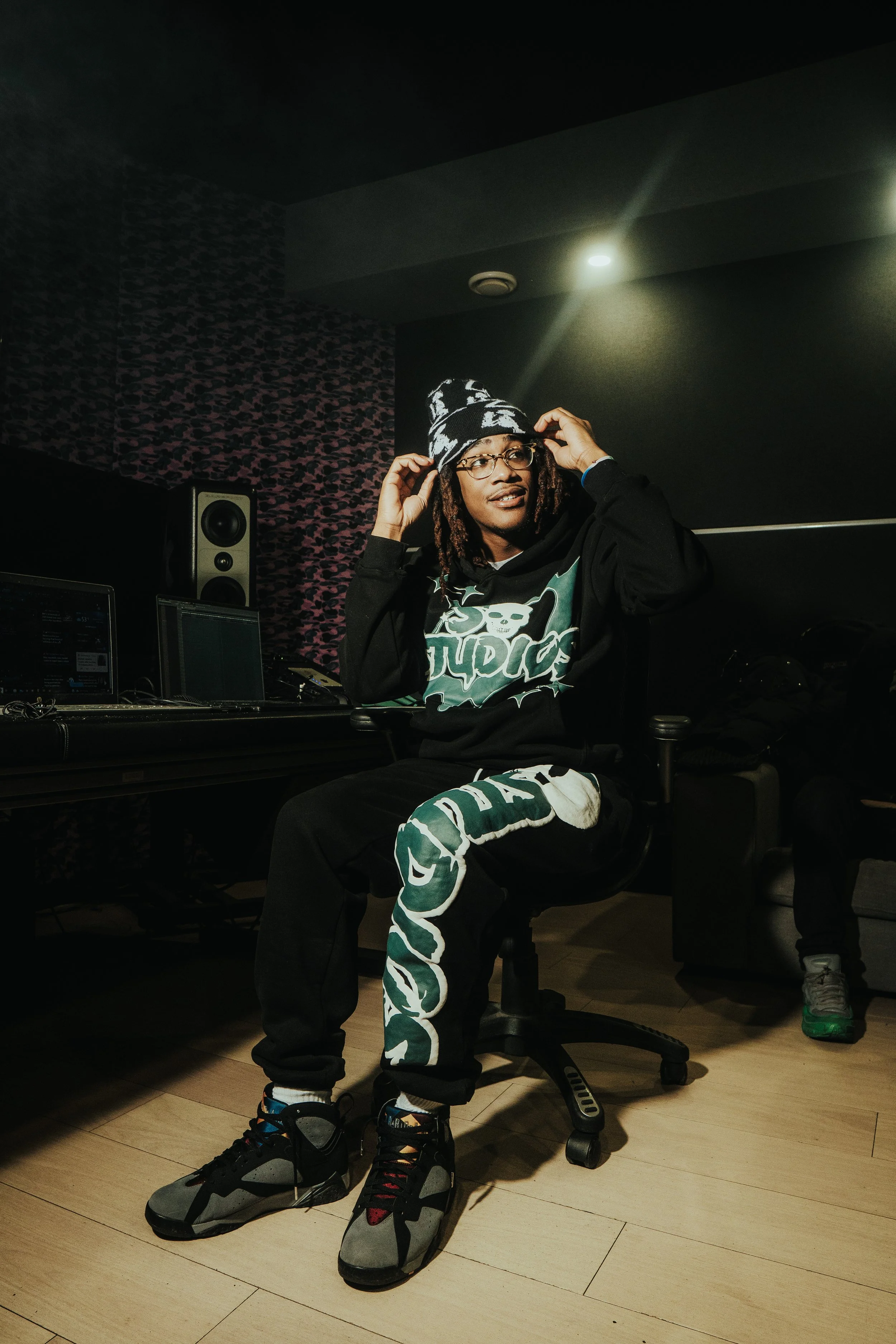 Young man with dreadlocks wearing glasses, a beanie, and stylish clothing sitting in a music studio.