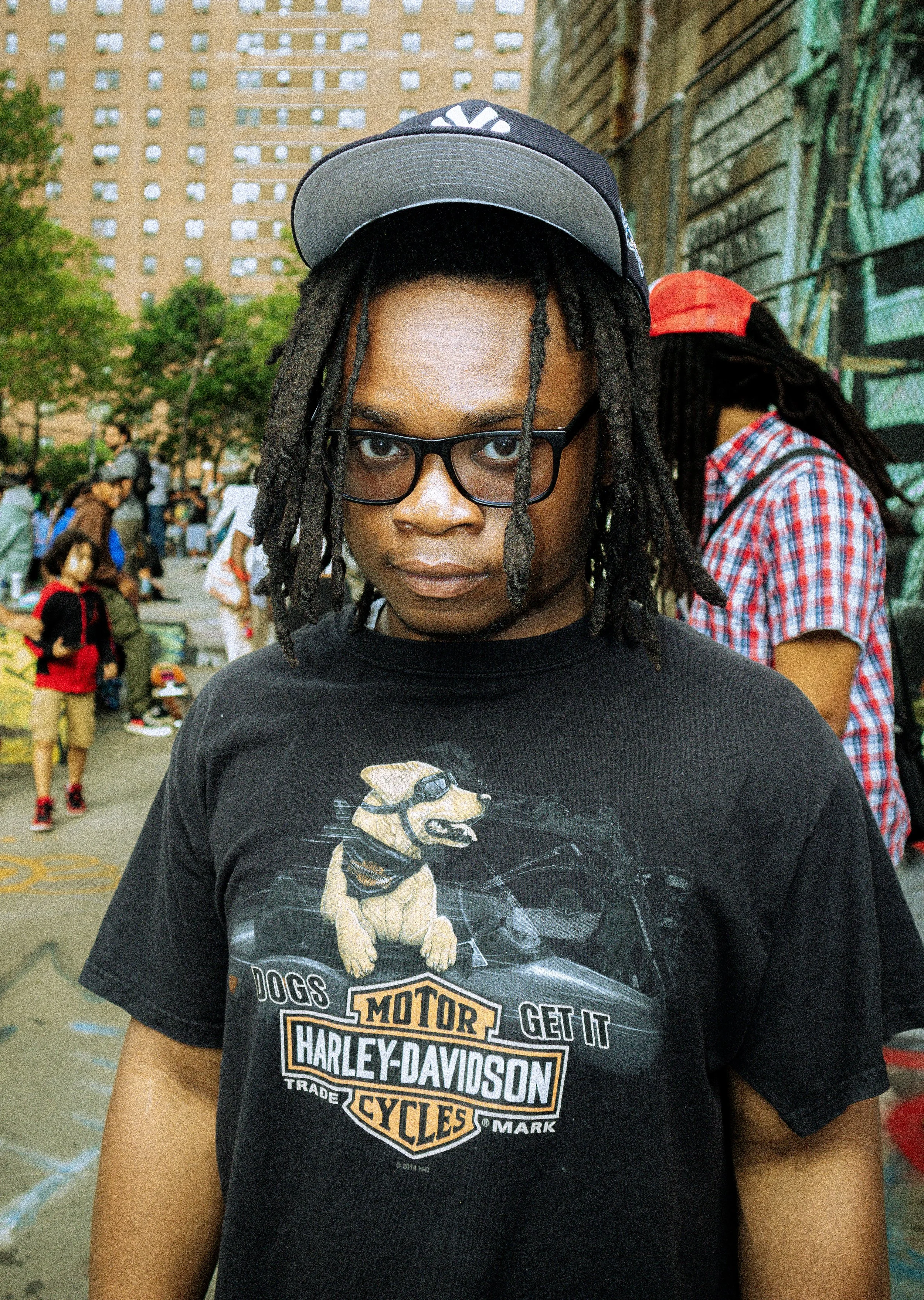 A young person with glasses and dreadlocks wearing a Harley-Davidson t-shirt and a baseball cap, standing outdoors in an urban park or plaza.