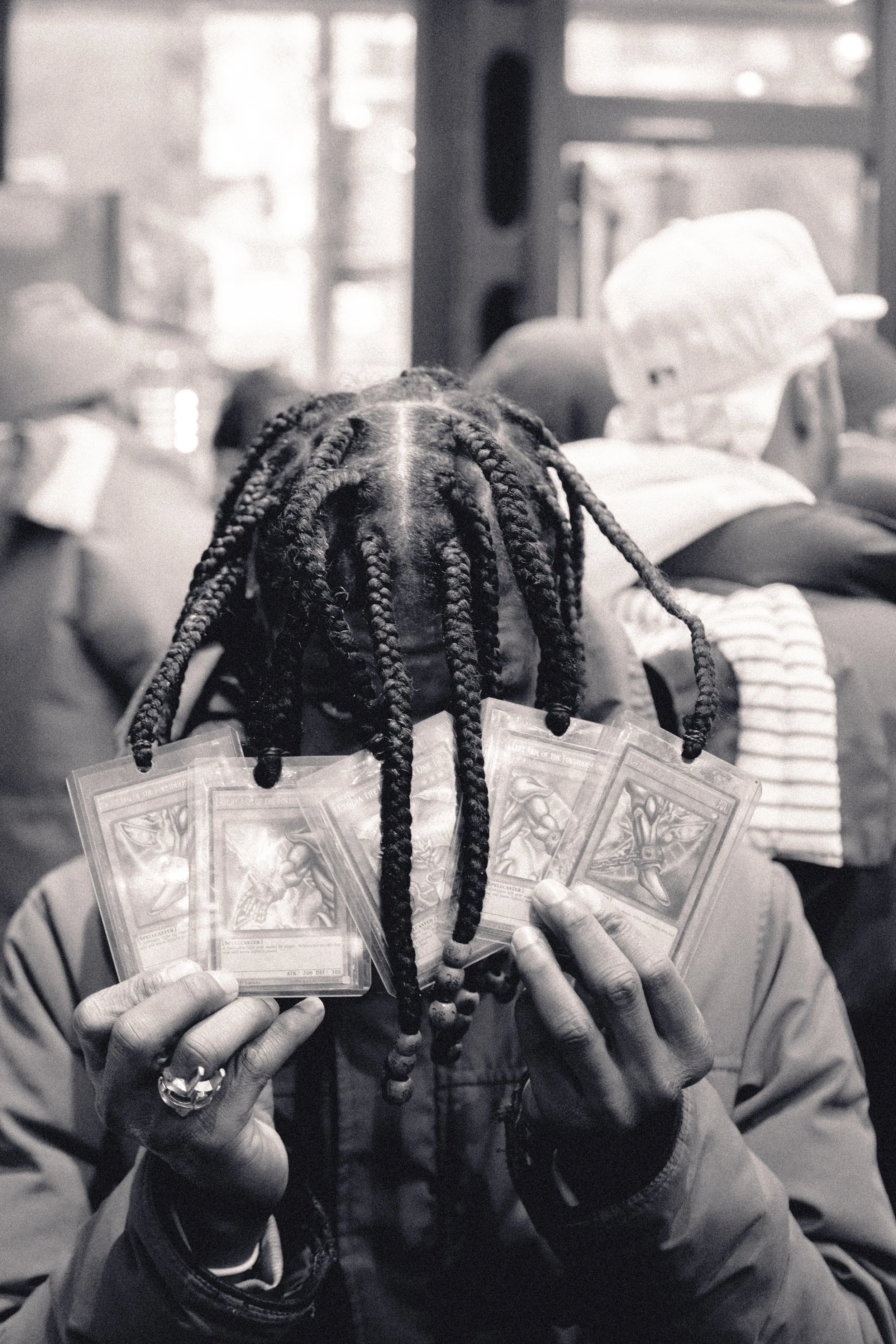 Person with braided hair holding multiple trading cards, standing in a crowded indoor space.