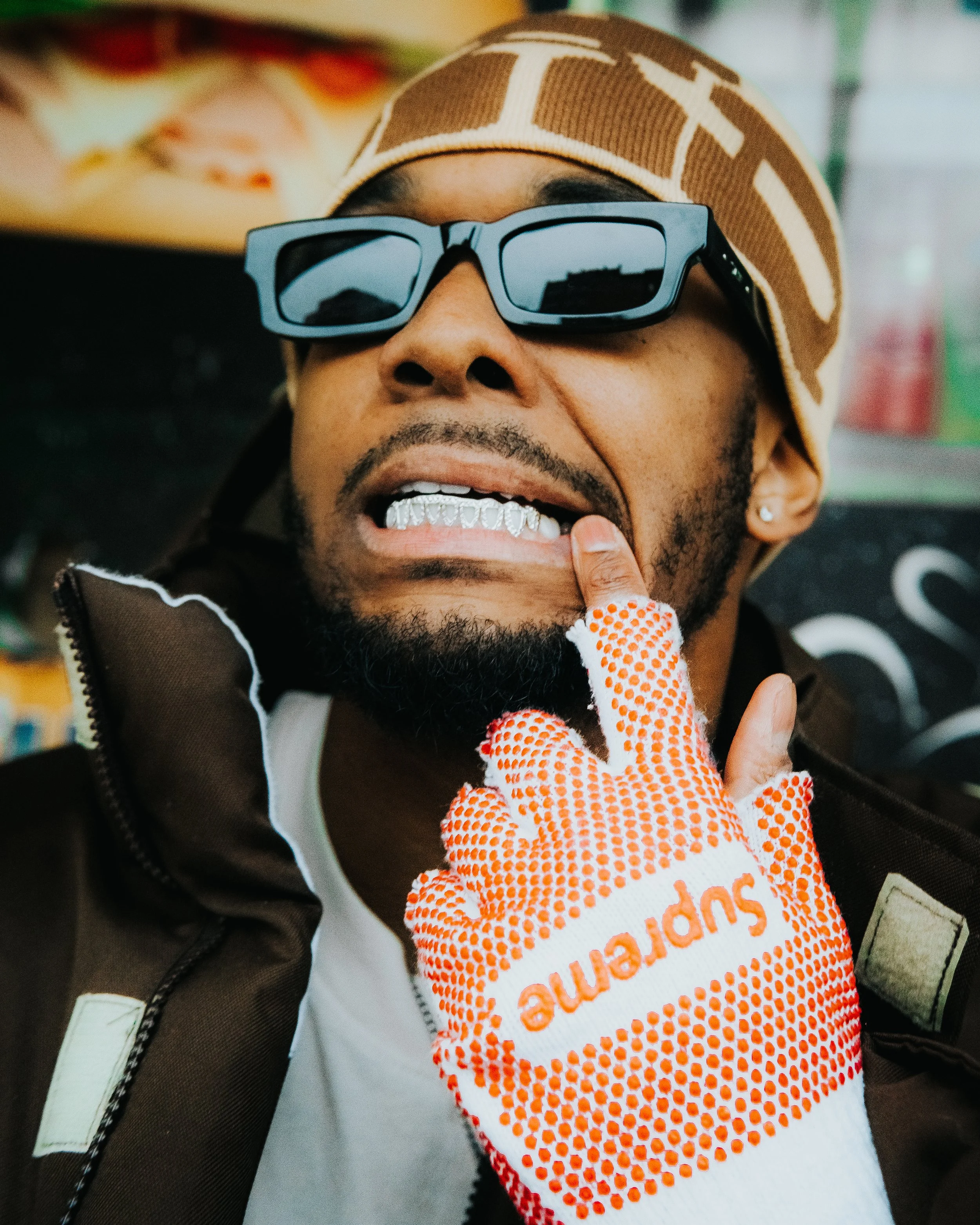 A man wearing sunglasses, a brown and beige hat, and a brown jacket with a white shirt, showing his dental jewelry, with an orange and white glove touching his lip in a close-up portrait.