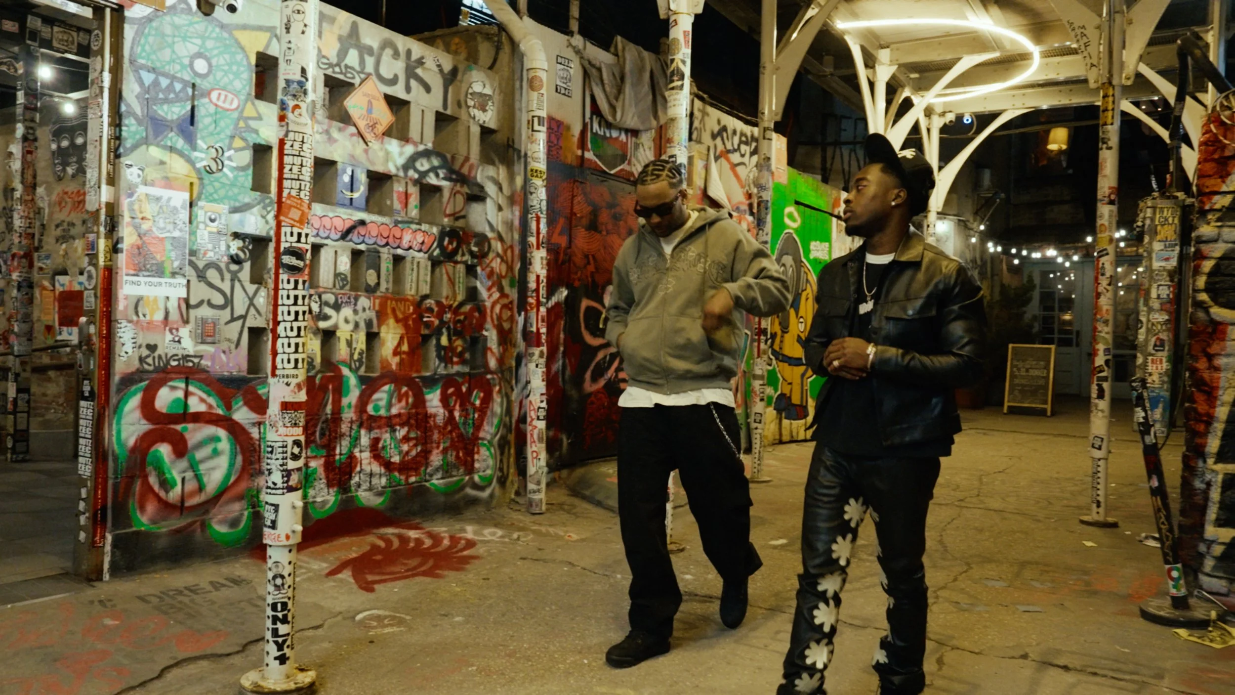Two people walking in a graffiti-covered alley with street art, stickers, and posters on walls and pipes.