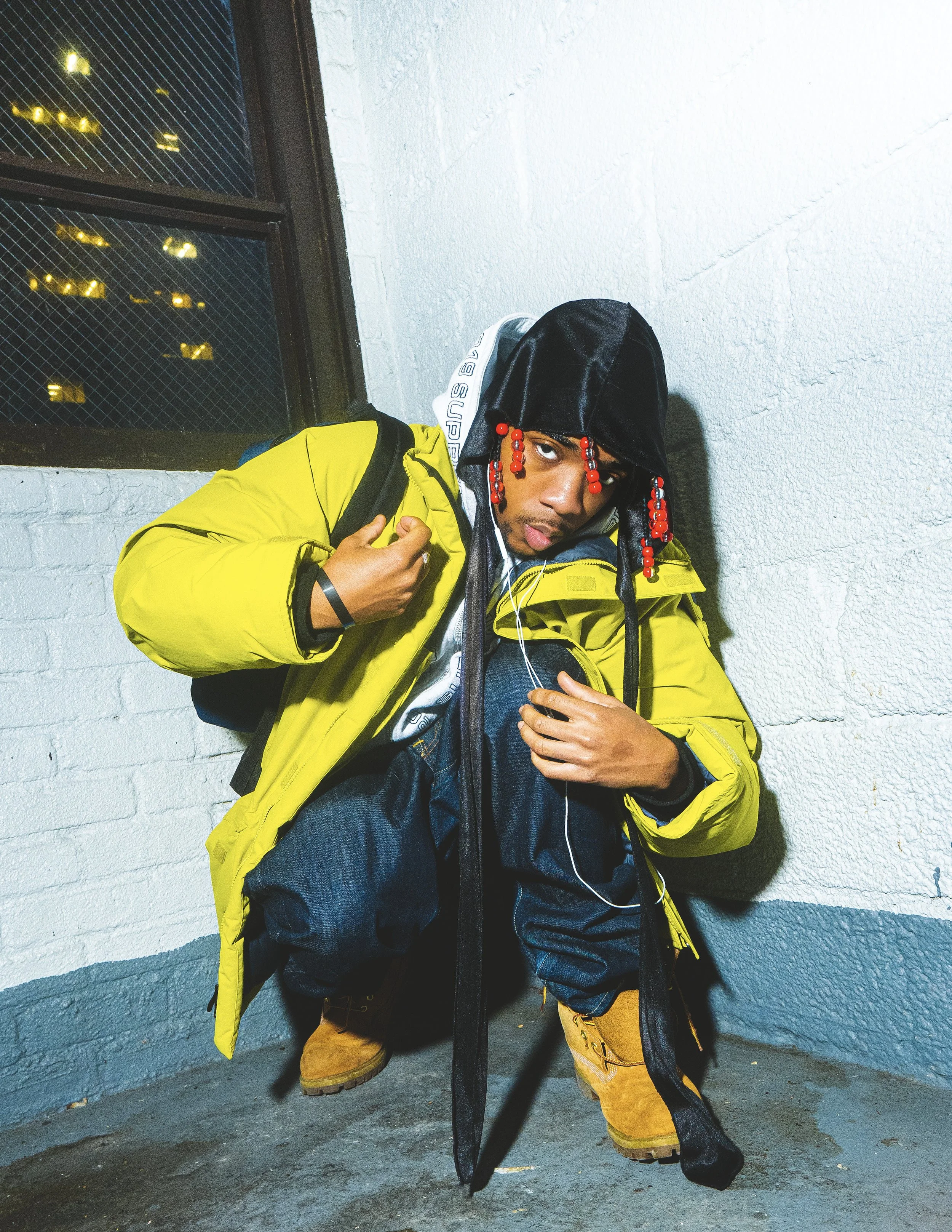 A young man crouching against a white textured wall next to a window with yellow lights. He is wearing a bright yellow jacket, black hood, yellow boots, jeans, and carries a backpack. He has red beaded hair accessories and is holding earphones with o