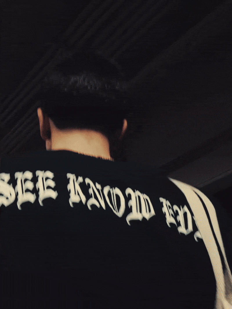 Back of a person wearing a black t-shirt with white text, and a black baseball cap, facing away in a dark indoor setting.