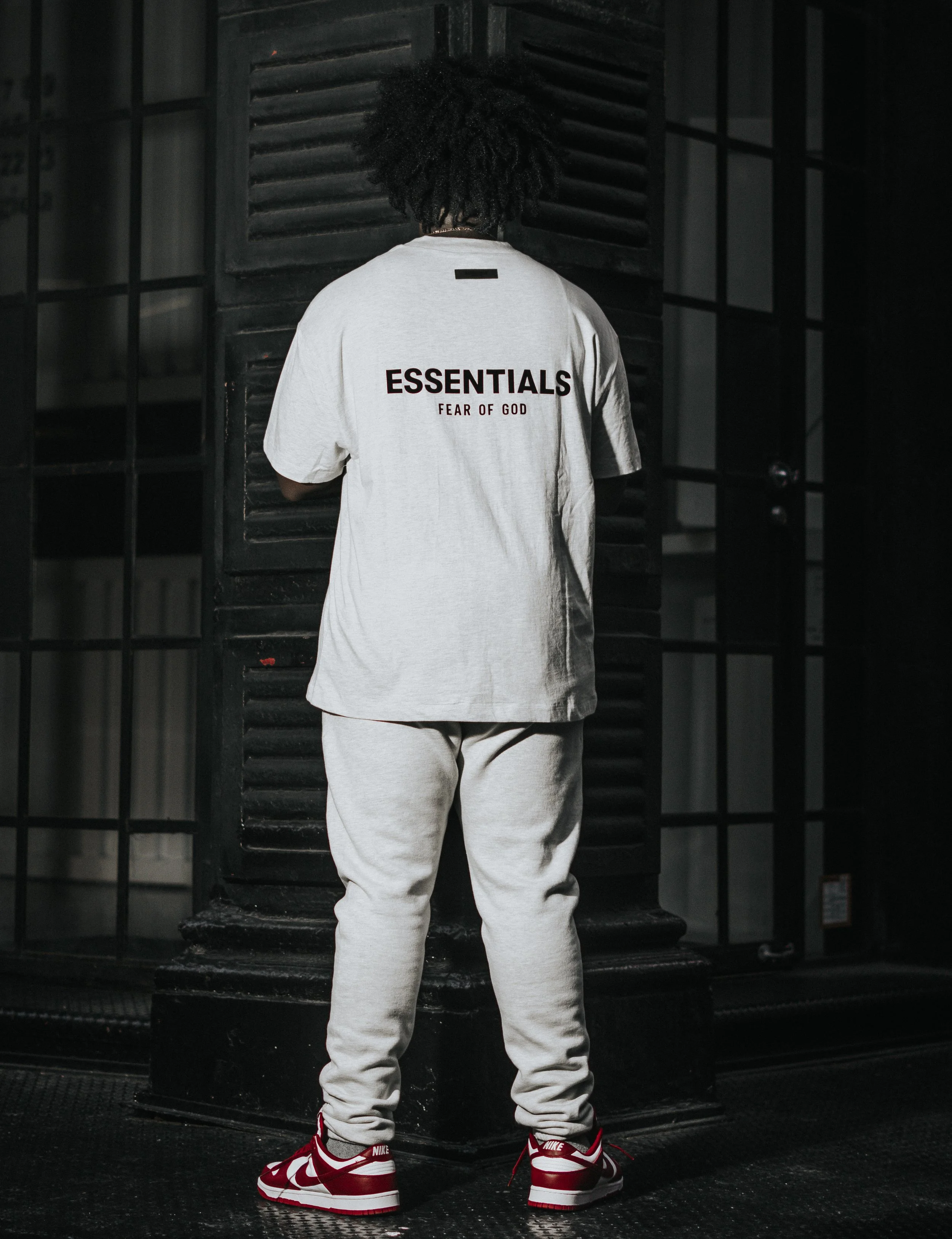 Person standing with back to camera, wearing a white T-shirt with 'ESSENTIALS FEAR OF GOD' printed on it, gray sweatpants, and red and white Nike sneakers, indoors near a dark pillar and black metal door.