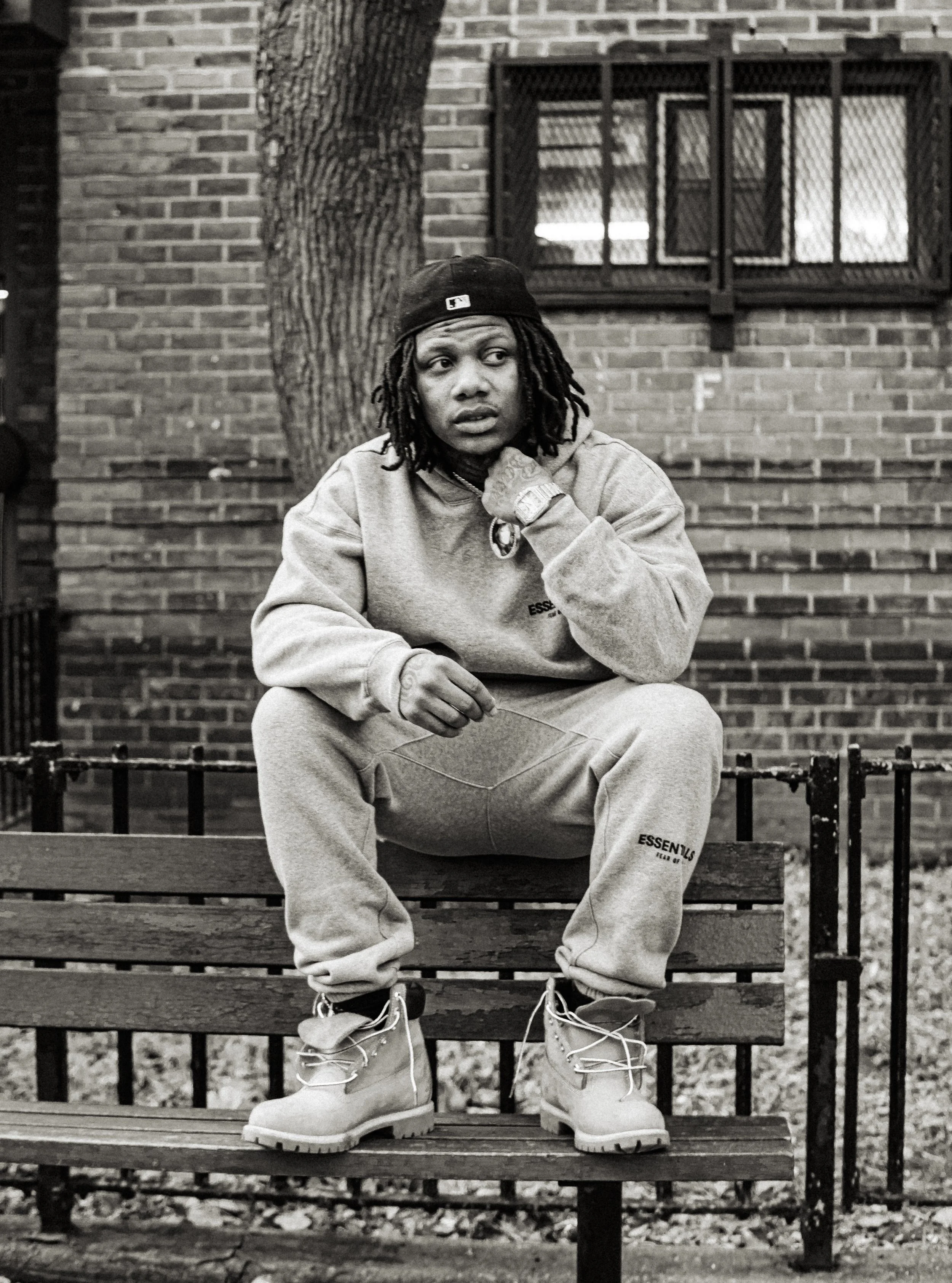 A person with dreadlocks sitting on a park bench, wearing a hoodie, sweatpants, and boots, with a serious expression, in front of a brick building and a tree.