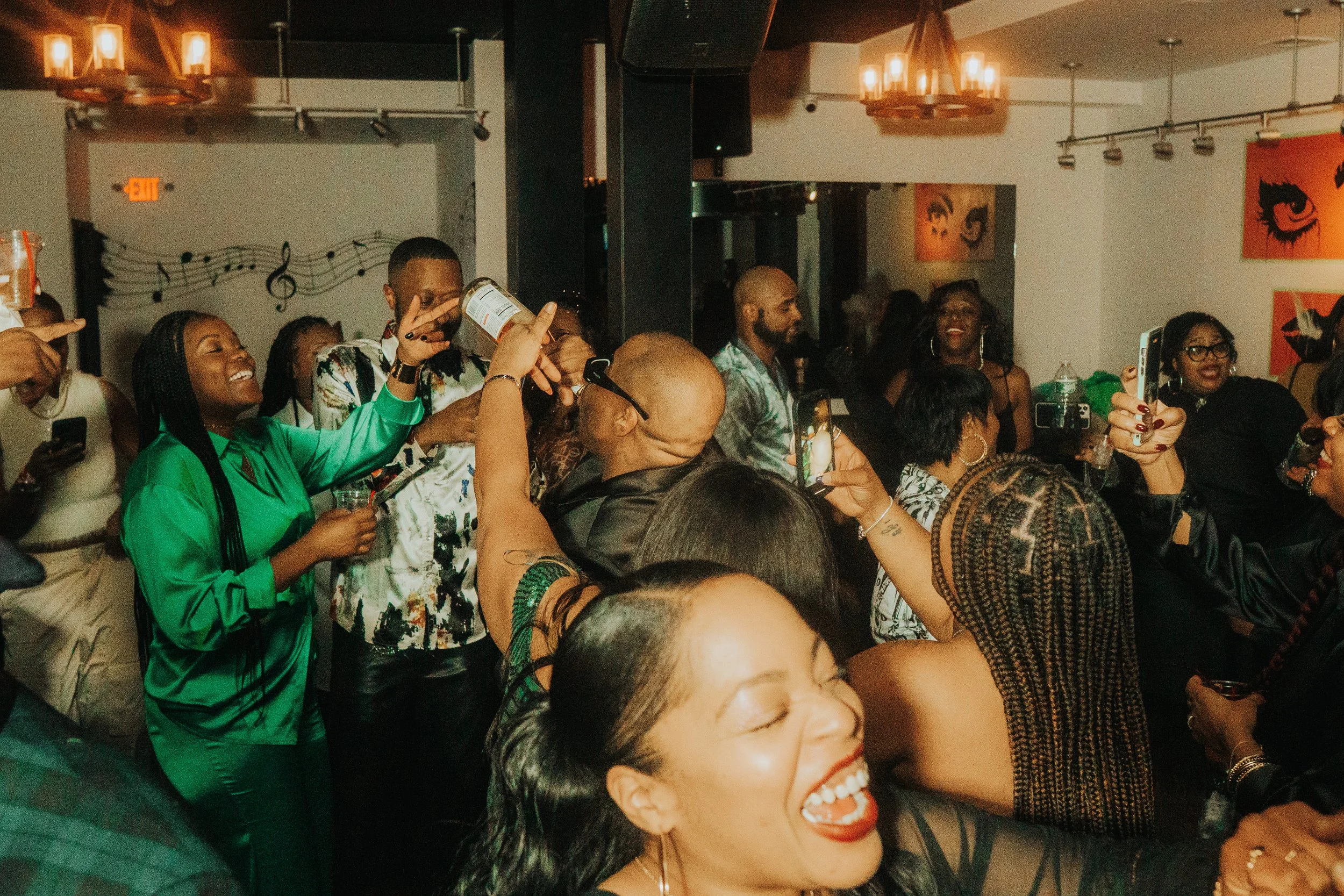 A lively indoor party scene with people enjoying music, dancing, and laughing. Attendees are holding drinks and capturing moments on their phones. The room is decorated with musical notes on the walls and warm lighting.