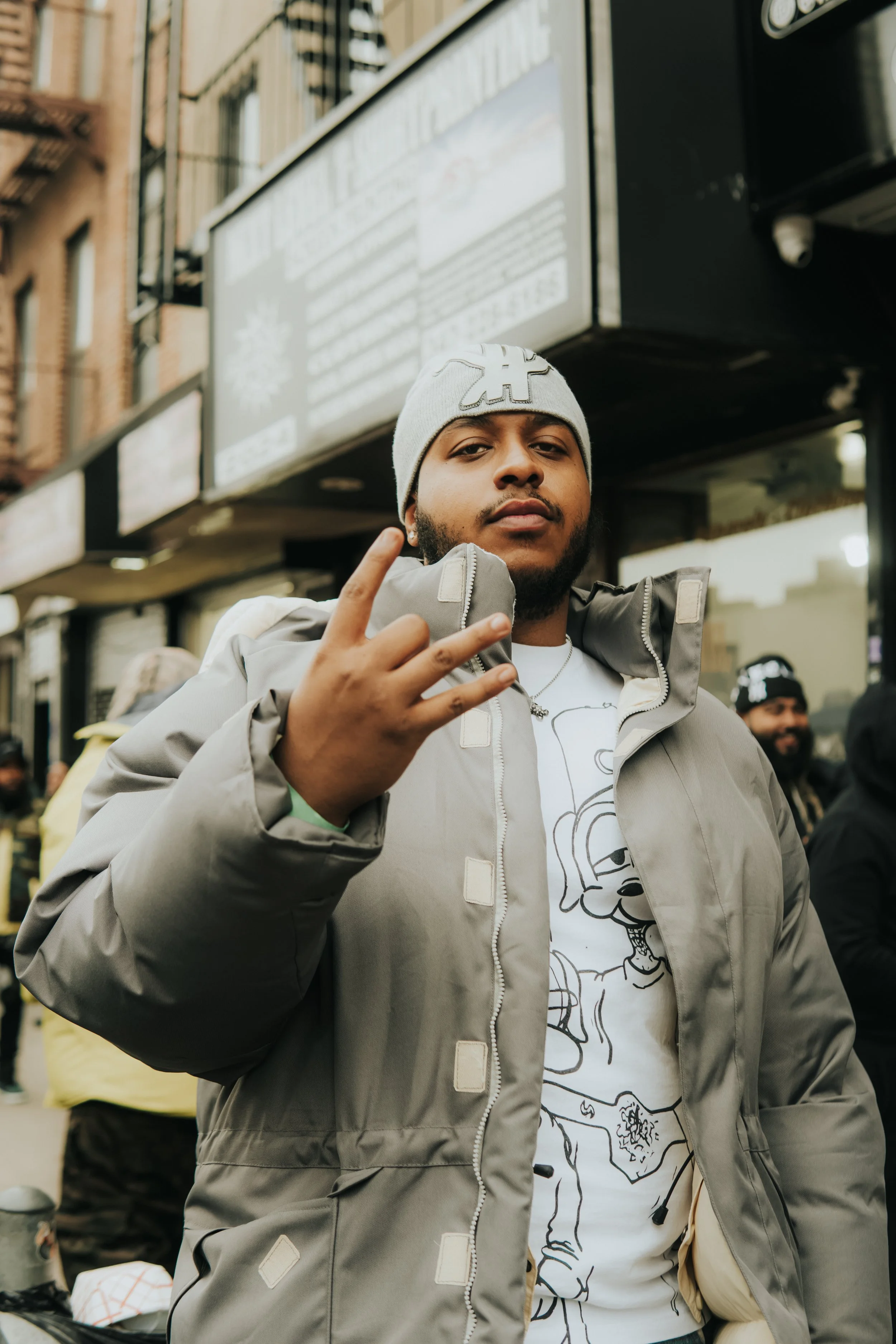 A young man standing on a city street, wearing a gray jacket, white graphic t-shirt, and gray beanie, making a hand gesture with his fingers, while people are in the background.