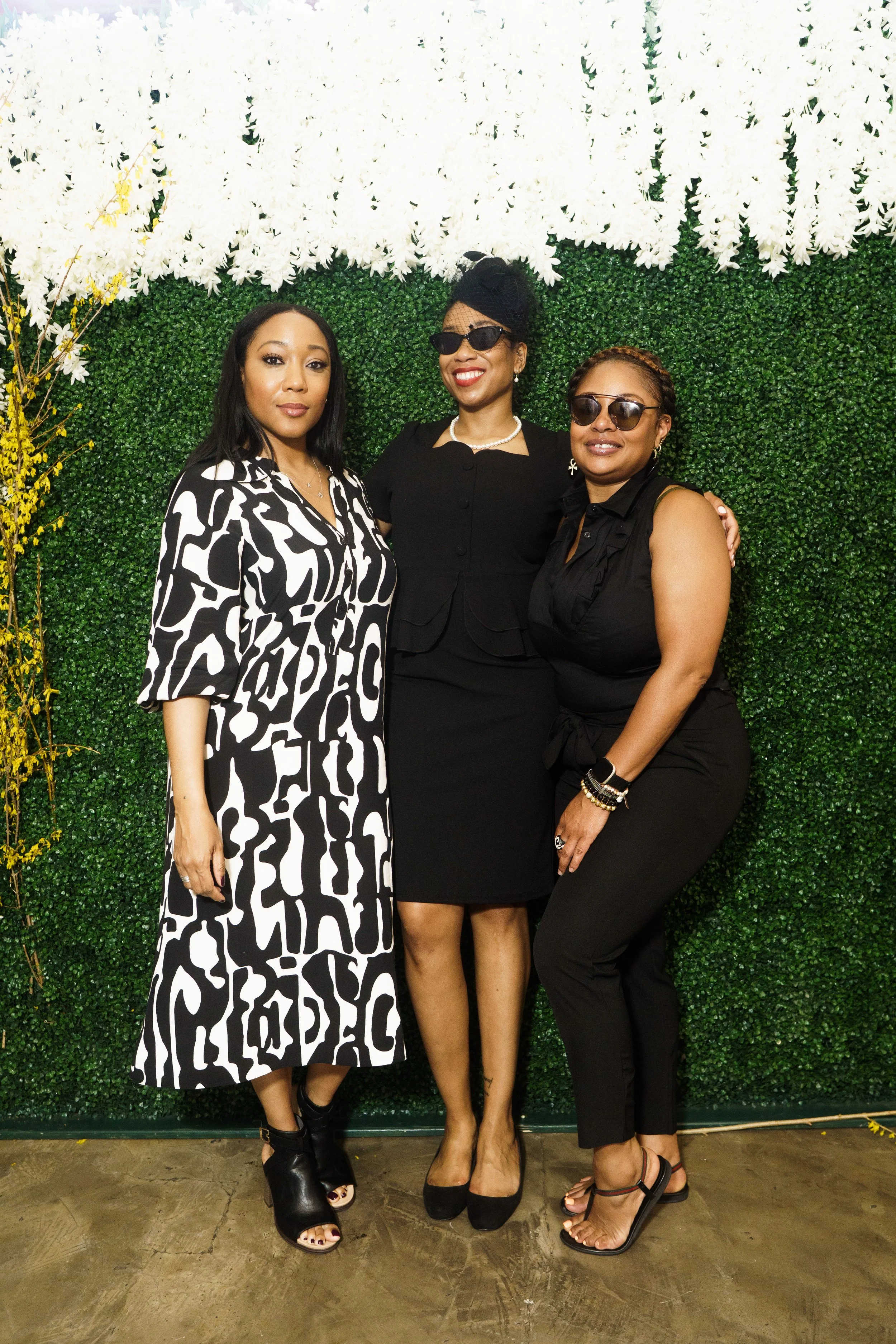 Three women posing in front of a green foliage wall; one wearing a black and white patterned dress, another in a black outfit with sunglasses, and the third in a black sleeveless top with sunglasses.