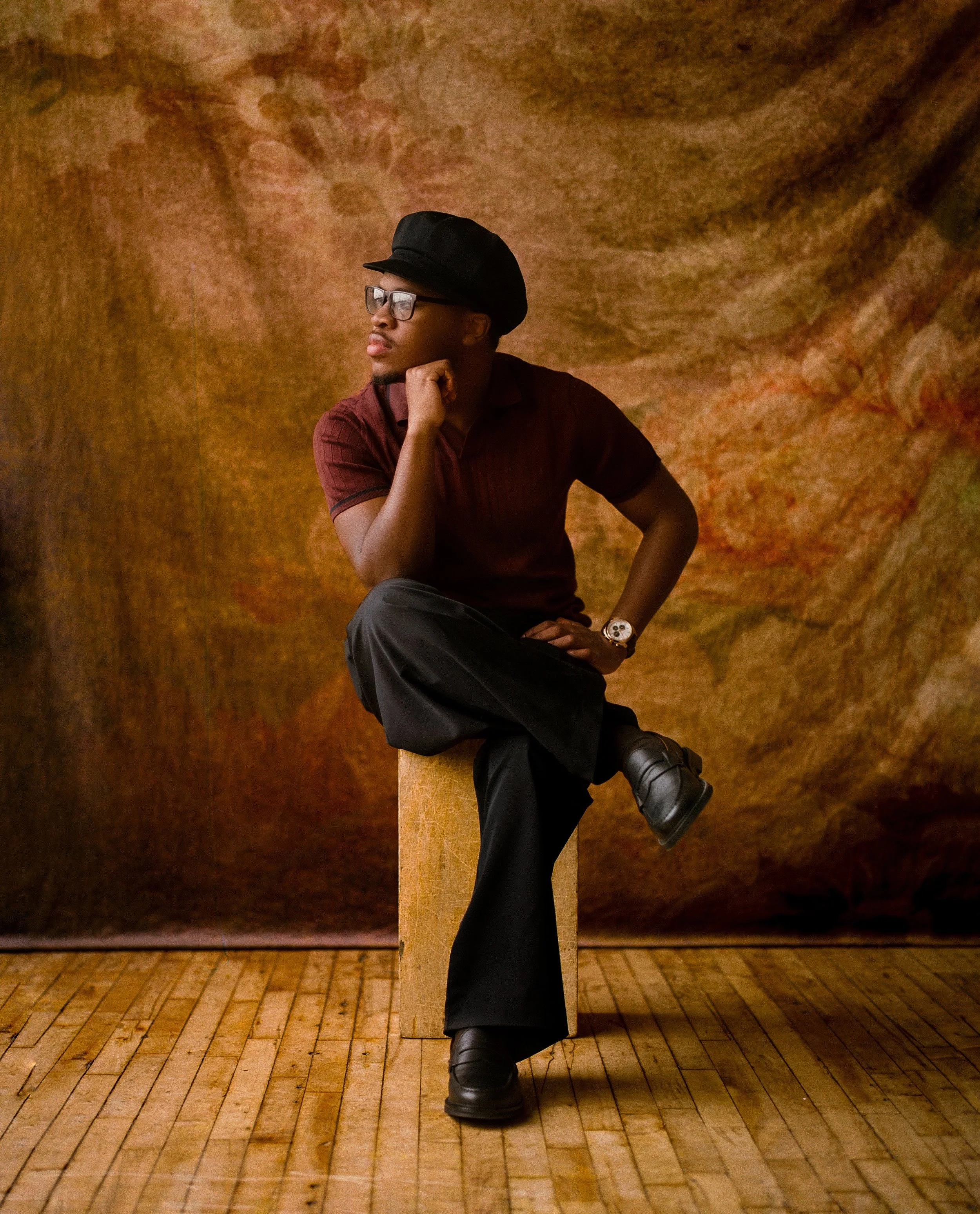 A man with glasses, a black hat, and a watch, wearing a maroon shirt and black pants, sitting on a wooden box with his legs crossed, resting his chin on his left hand, against a textured brown and orange background.