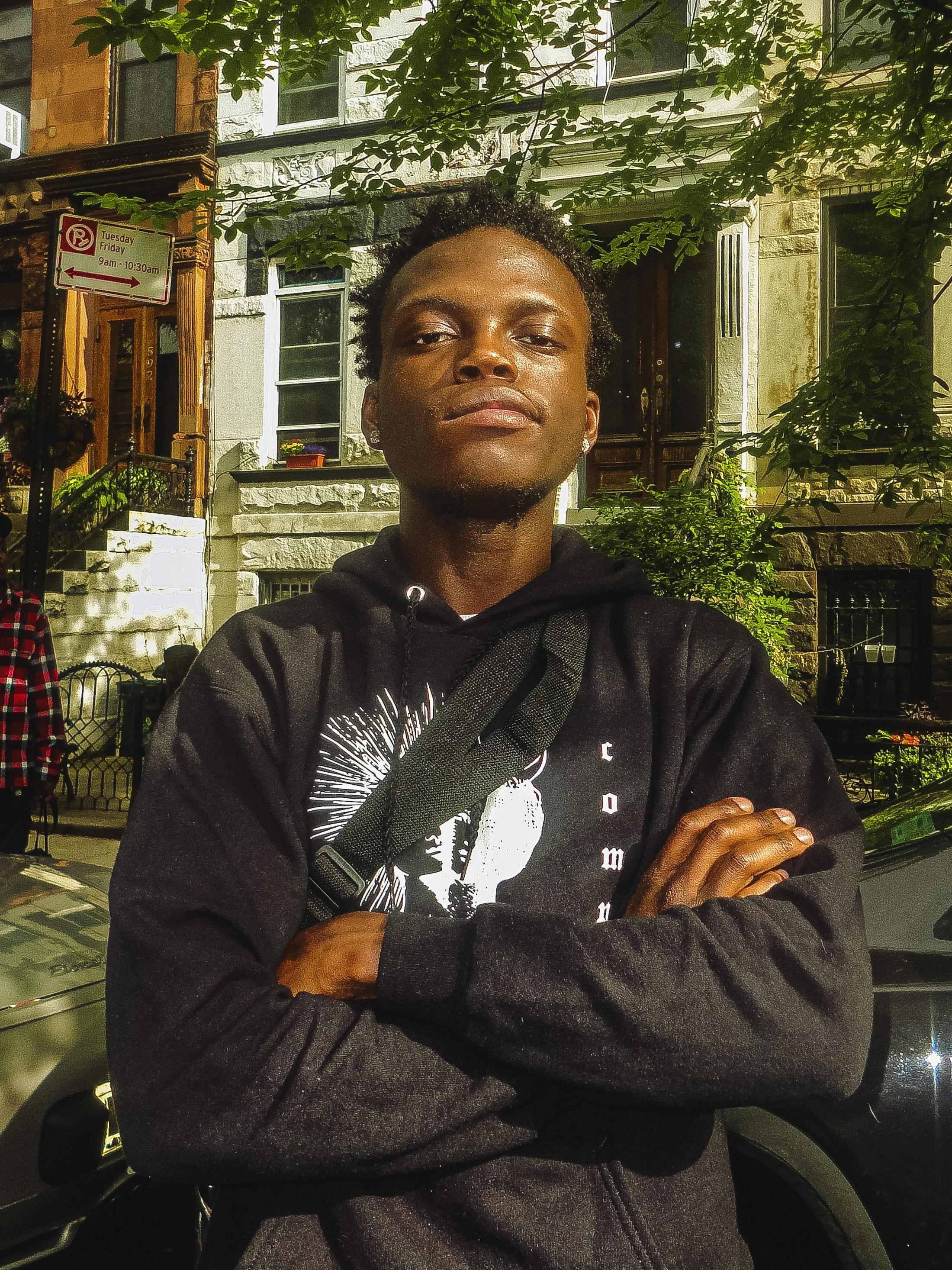 Young man with arms crossed standing on a city street at night, wearing a black hoodie with graphic design, in front of residential buildings with stairs and greenery.