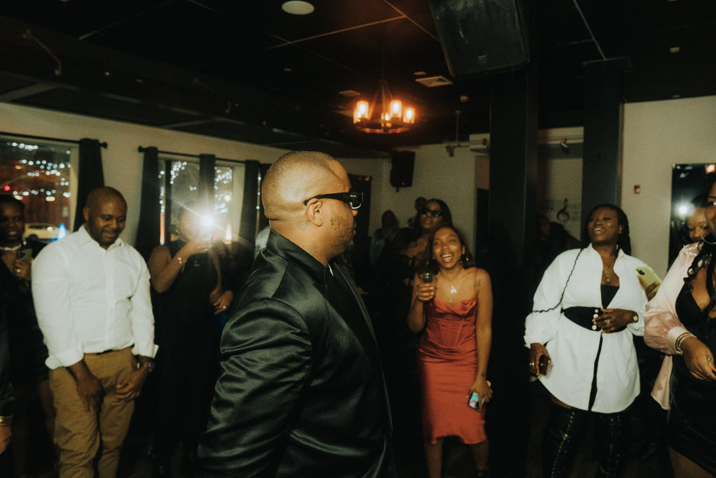 Group of people at a party, some smiling, with the man in the foreground wearing sunglasses and a dark suit. The party has dim lighting with string lights visible through the windows. A woman in a red dress holds a drink while another in white smiles