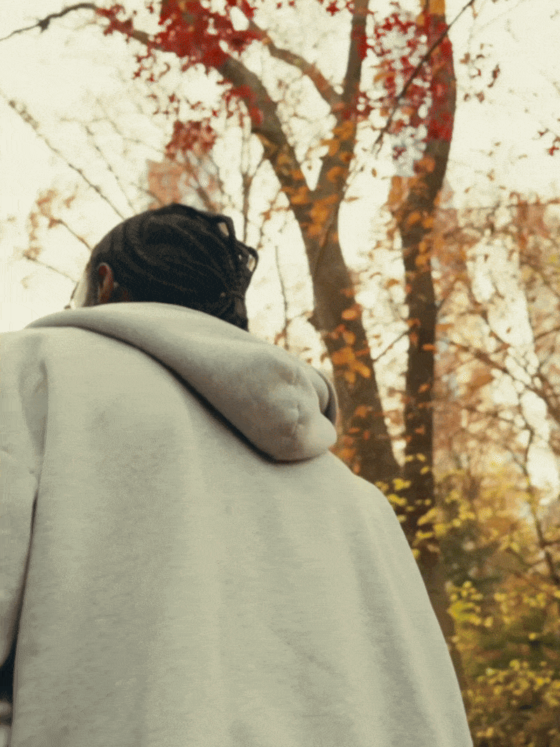 Back view of a person with dreadlocks wearing a light gray hoodie, standing outdoors with autumn-colored trees in the background.