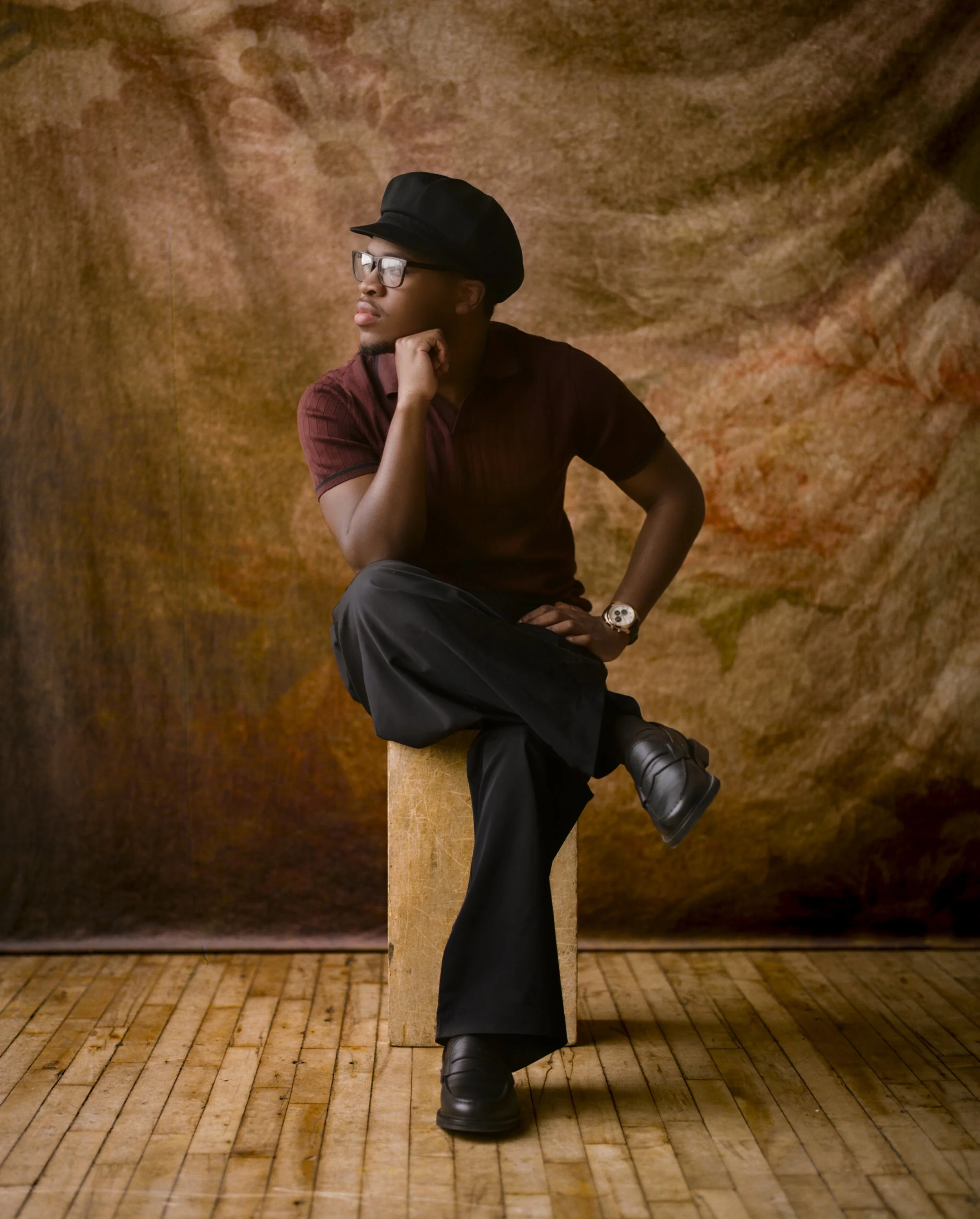 A man sitting on a wooden block against a textured brown background, wearing a black hat, glasses, a short-sleeved maroon shirt, black pants, and black shoes, with his right hand resting on his chin and his left hand on his knee.