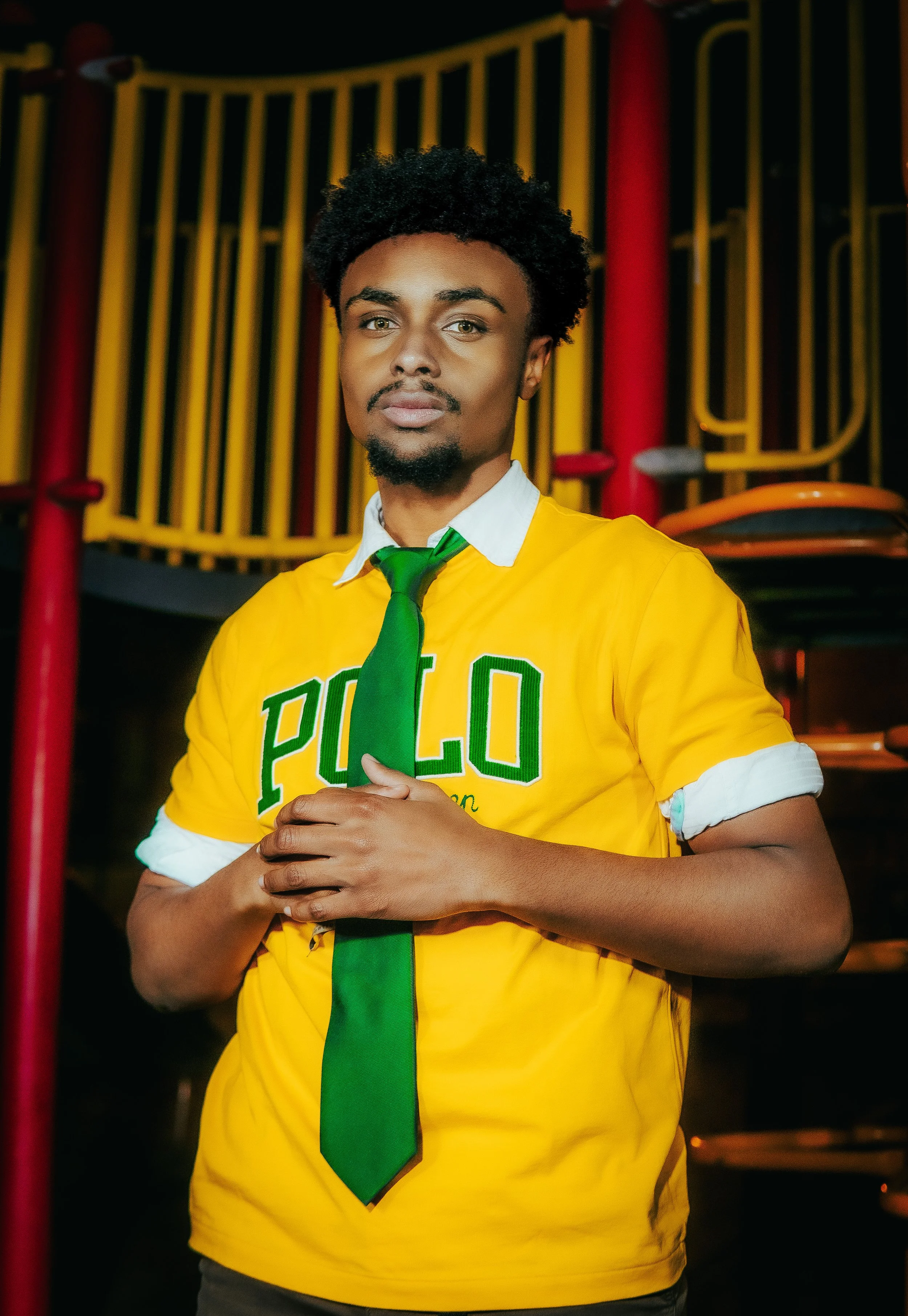 A young man with curly black hair and a goatee wearing a yellow Polo shirt with rolled-up sleeves, a white collar, and a green tie standing indoors with a colorful playground structure in the background.