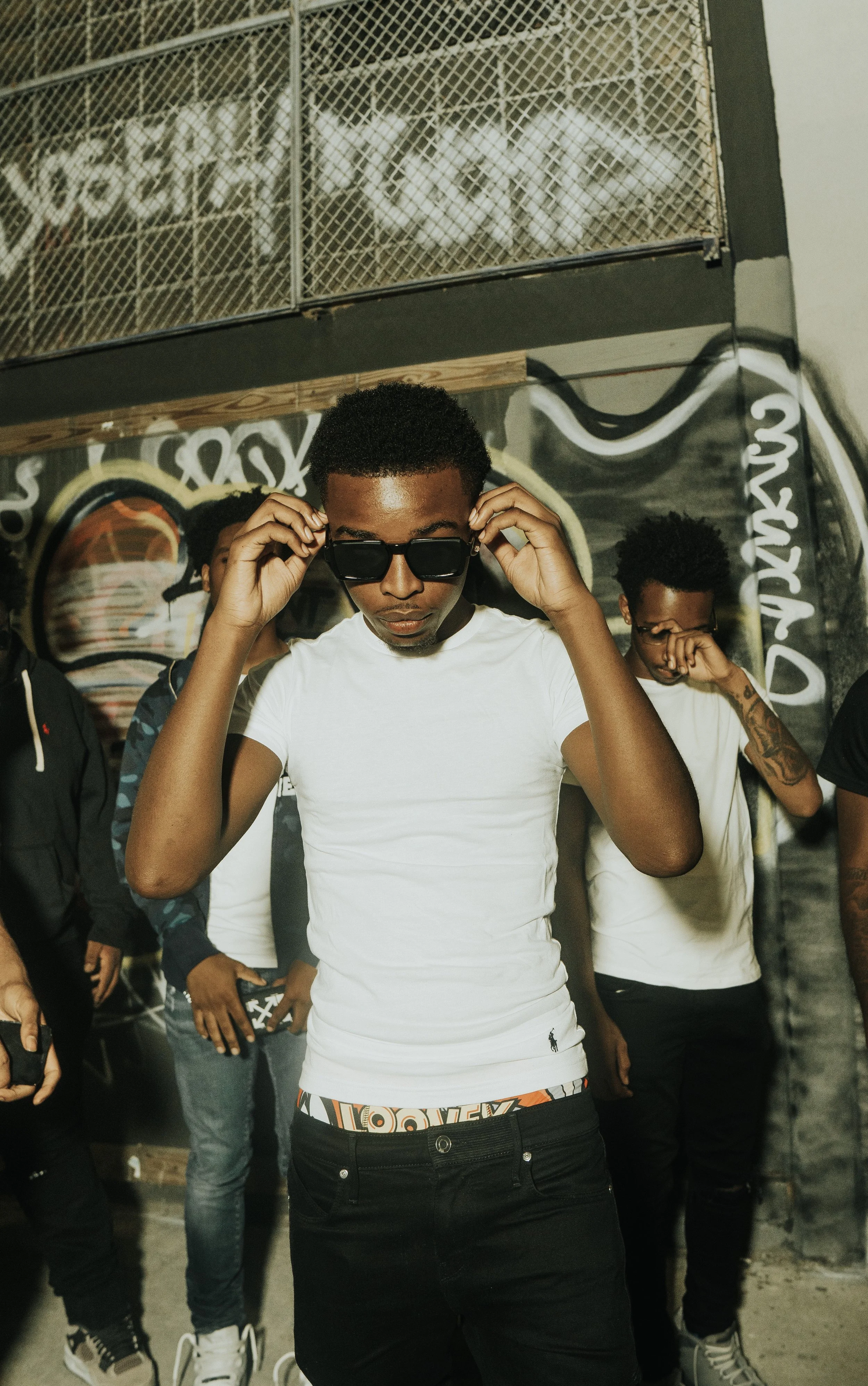 A young man wearing sunglasses and a white T-shirt adjusting his glasses in an urban setting with graffiti art and other people behind him.