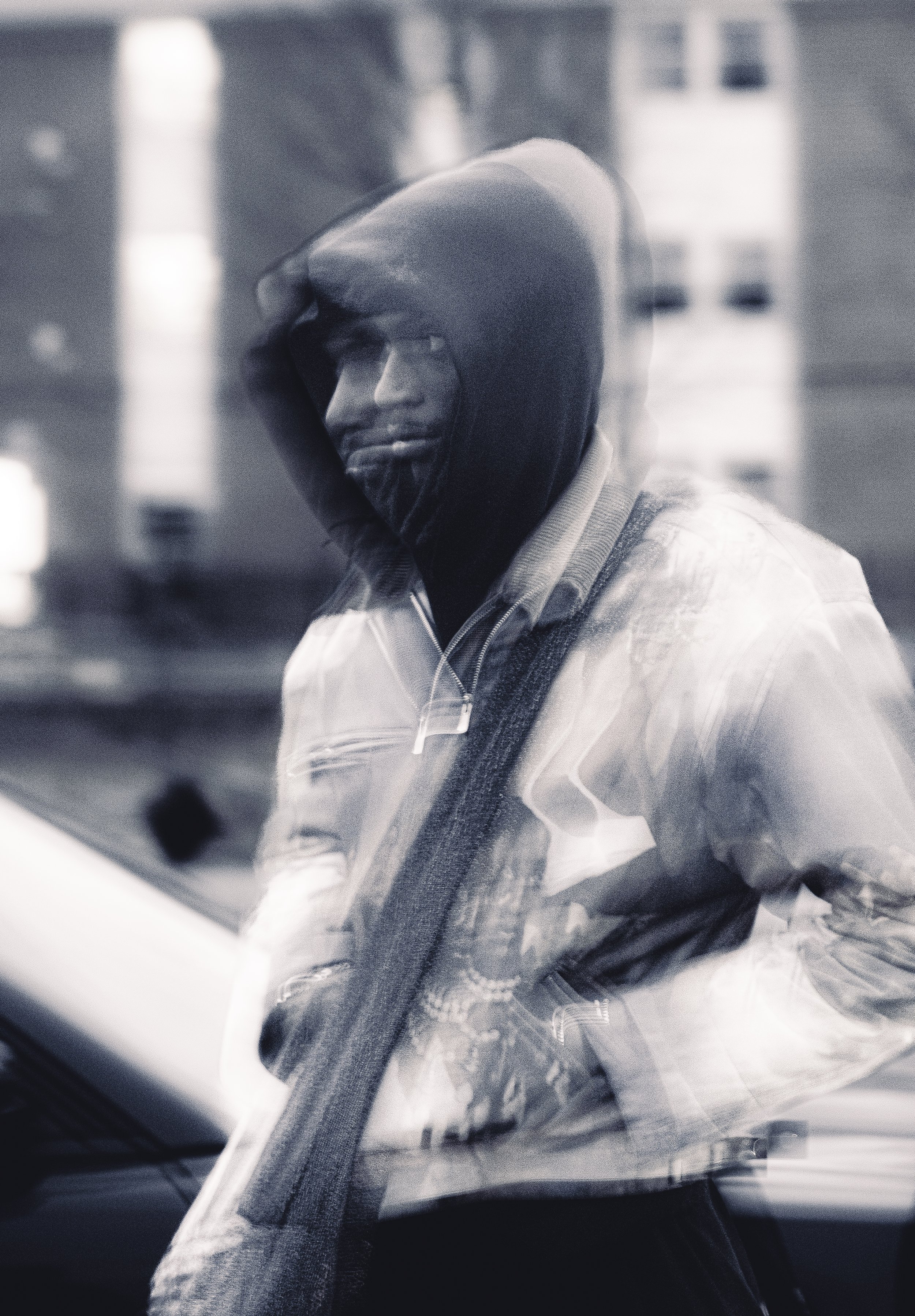 A person wearing a hoodie, glasses, and a jacket, standing outdoors in an urban setting, seen through a window or glass surface with reflections.